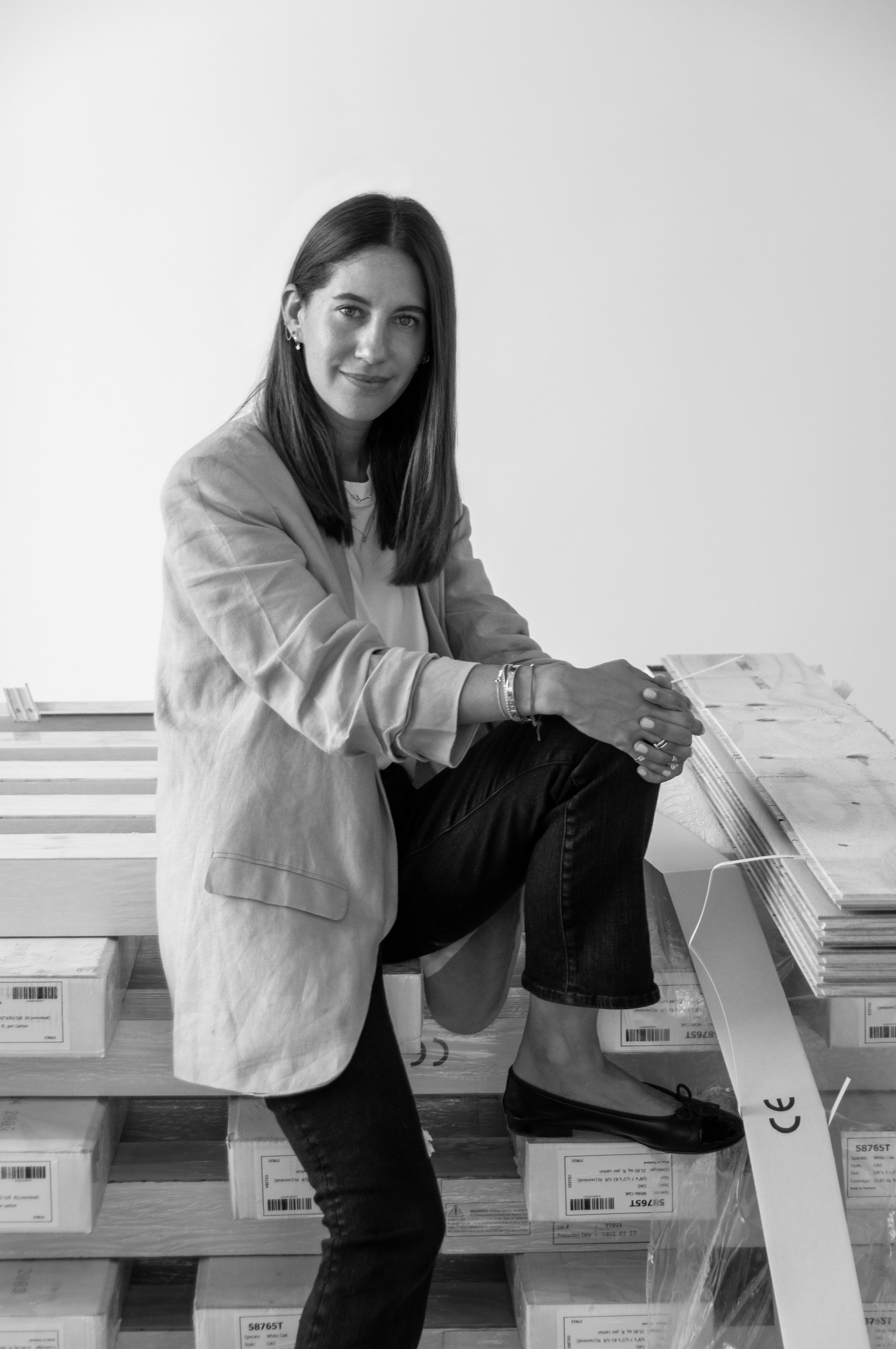 Black and white photo of a woman sitting on wooden pallets in what appears to be a warehouse or storage room, holding a box of white paper, with more boxes stacked behind her.