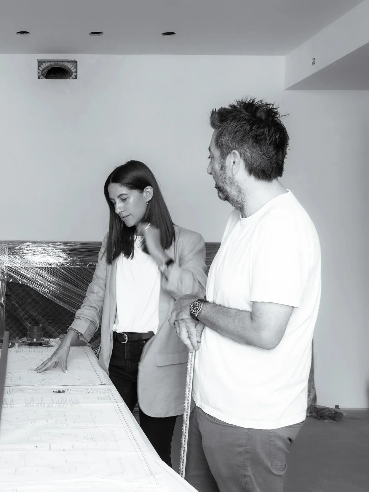 Two people, a woman and a man, are standing and looking at architectural plans or drawings on a table. The woman has straight dark hair, is wearing a light jacket and a white shirt, and is touching the plans. The man has short, slightly messy hair, a beard, and is wearing a white shirt with a watch on his left wrist. They are in a room with a plain wall, some ceiling lights, and a vent, possibly under construction or design planning.