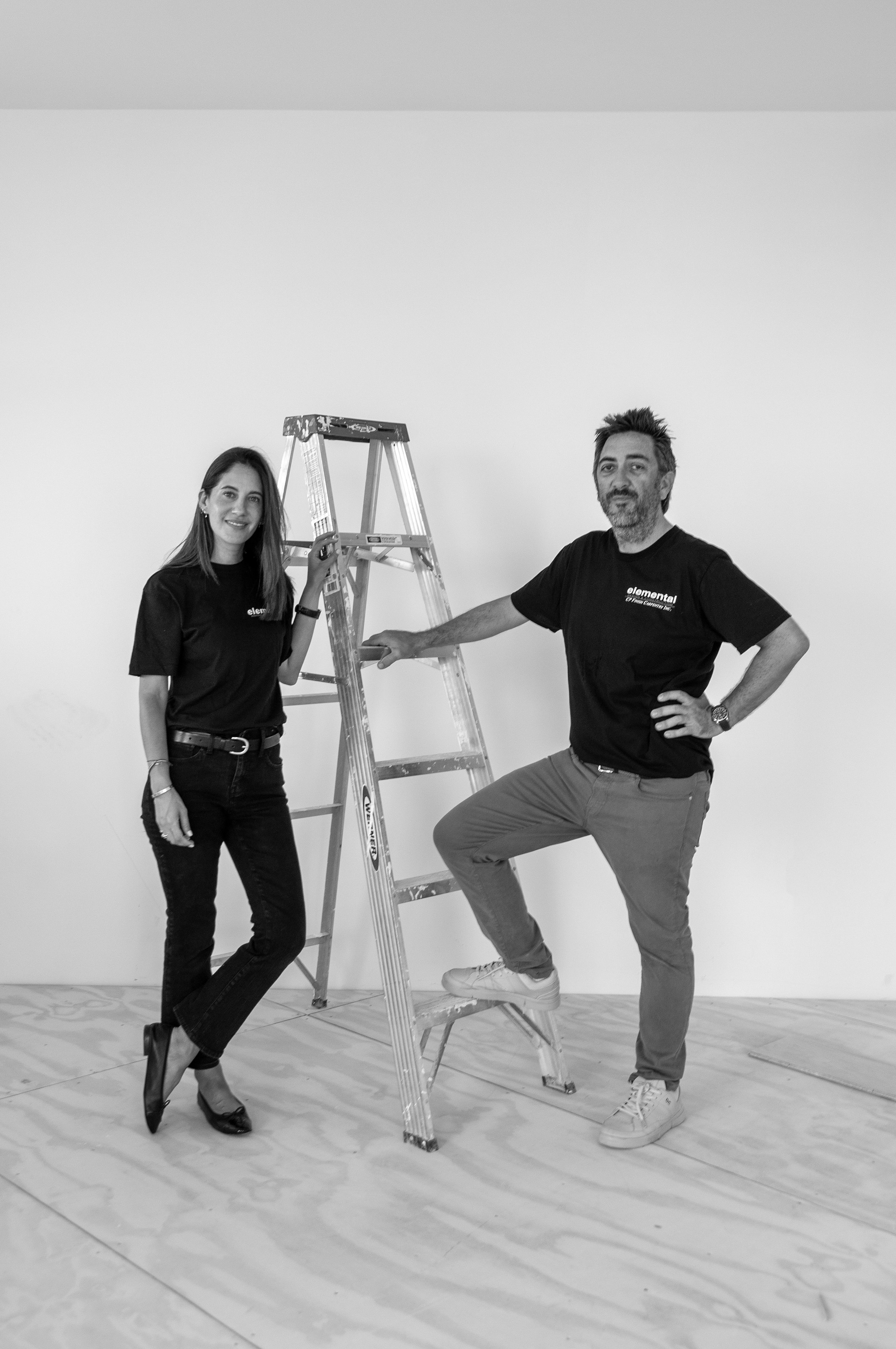 A woman and a man standing next to a ladder in a room with a plain wall and a floor with wooden pattern. The woman is dressed in black shoes and black pants, and the man is wearing casual clothes with sneakers. They are posing confidently with one foot on the ladder and smiling.