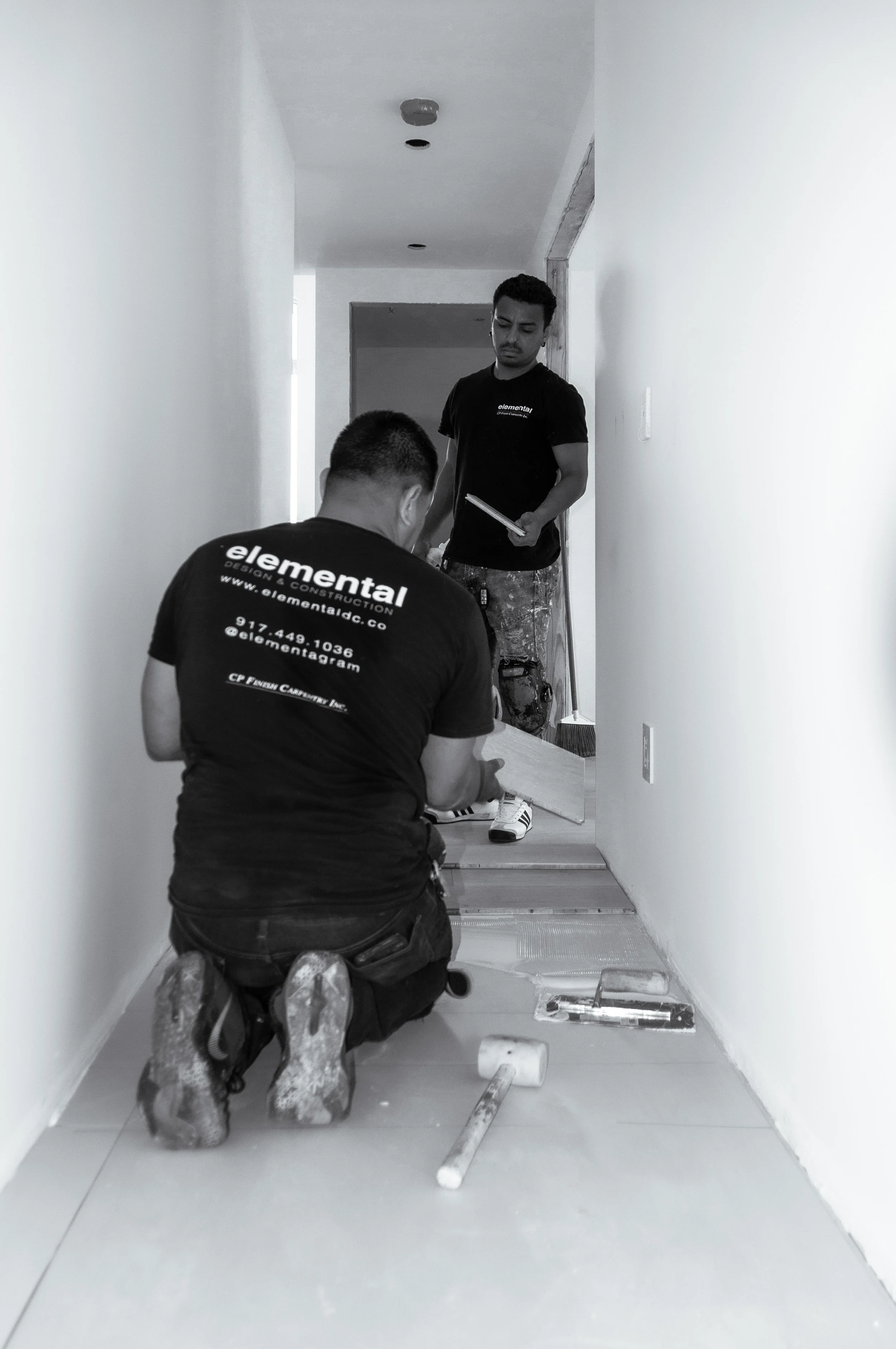Two workers installing or repairing a hallway floor, one kneeling and one standing, surrounded by tools and materials.