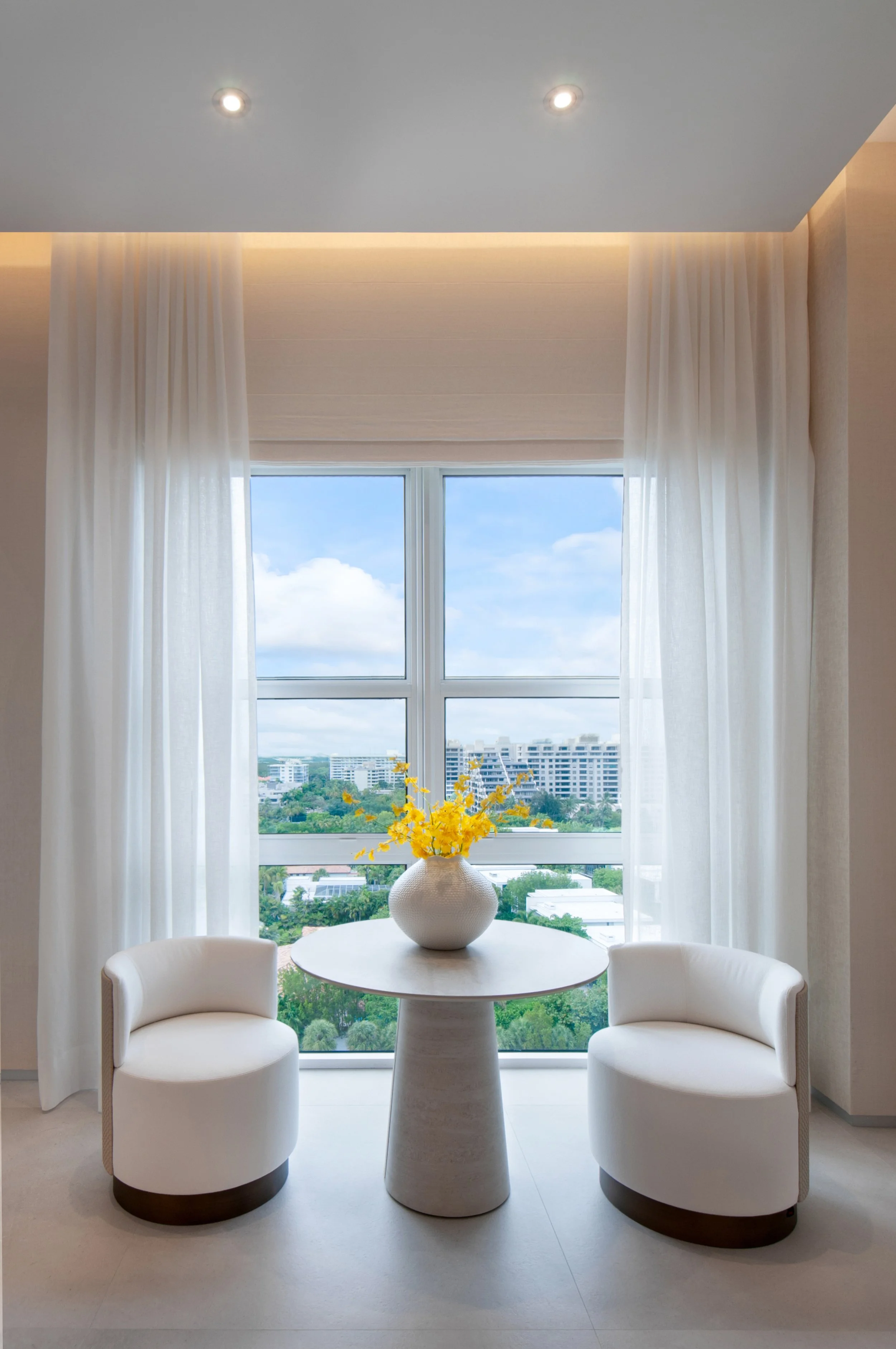 A bright, modern dining area with a round white table, two white curved armchairs, and a large window with sheer white curtains. A white vase containing yellow flowers is centered on the table. Outside the window, a cityscape with apartment buildings and lush greenery is visible, and the room has a high ceiling with recessed lighting.