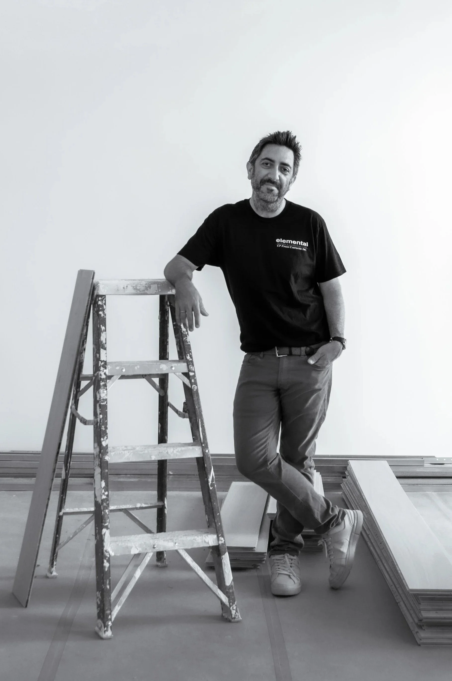 A man with dark hair and beard leaning on a ladder, standing next to a stack of tiles or panels in an unfinished room with a plain white wall.
