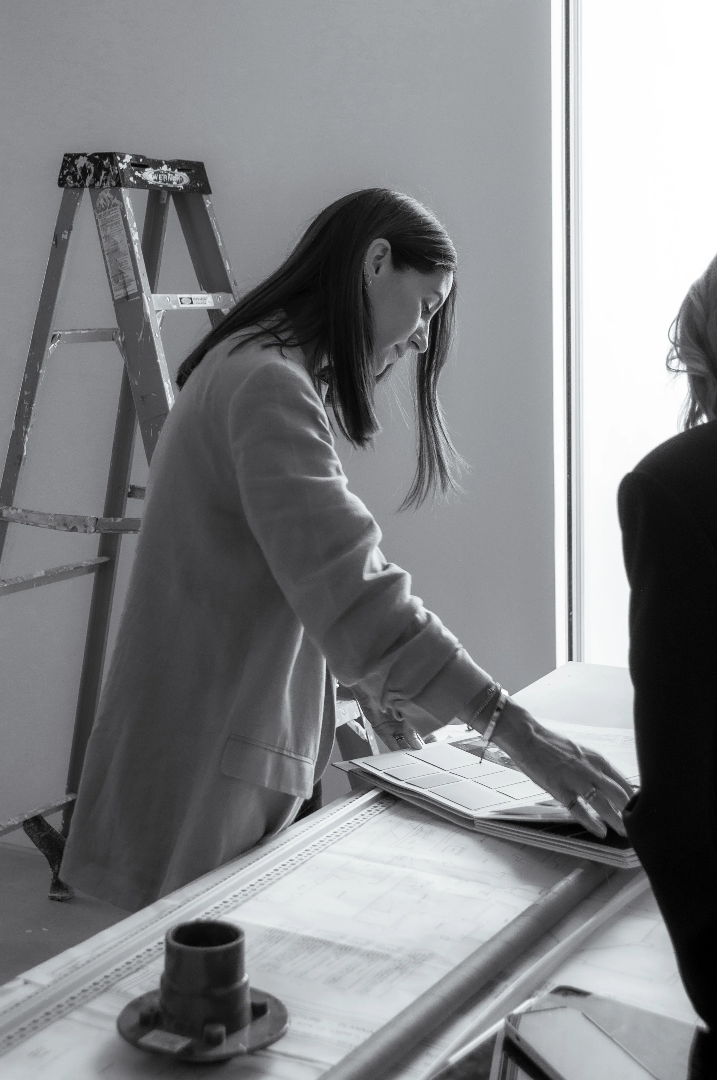 A woman with long hair, wearing a light-colored blazer, is looking down at architectural plans on a table. There is a ladder in the background and a window letting in natural light.