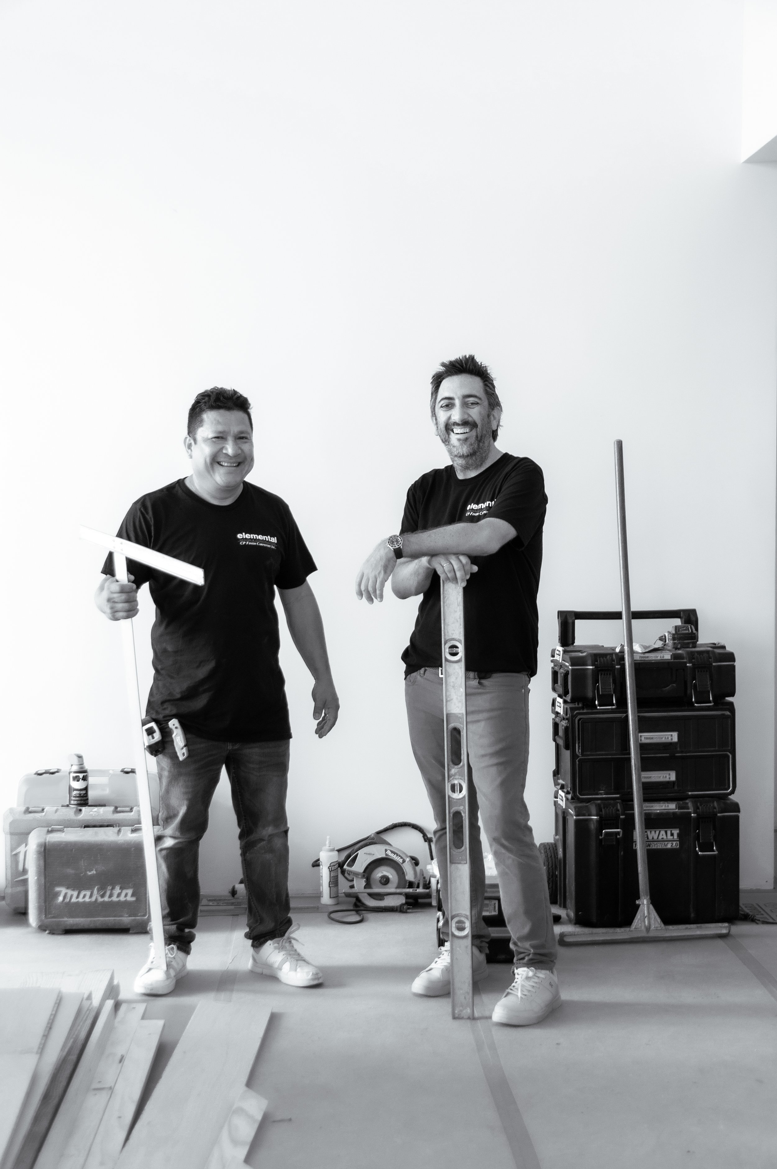 Two men standing in a room with construction tools, one holding a large level and the other resting on a small stepladder, both smiling.