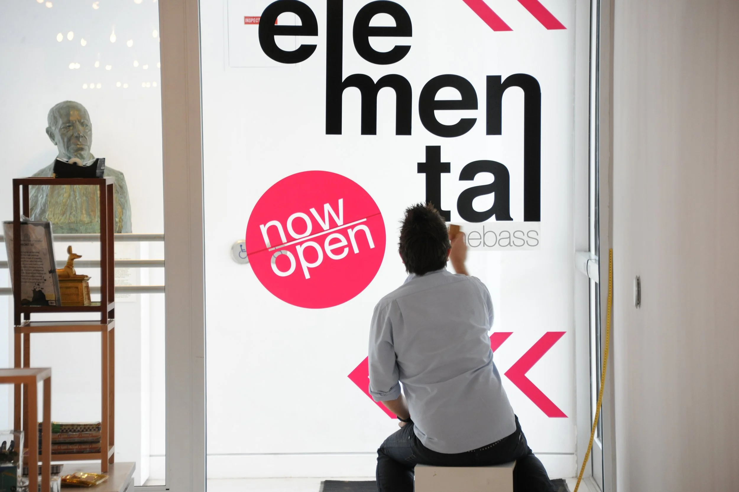 A man kneeling and placing a sticker on the glass door of a store with a sign that reads 'Edelmen' in large black letters, and a red circle with white words 'now open'.
