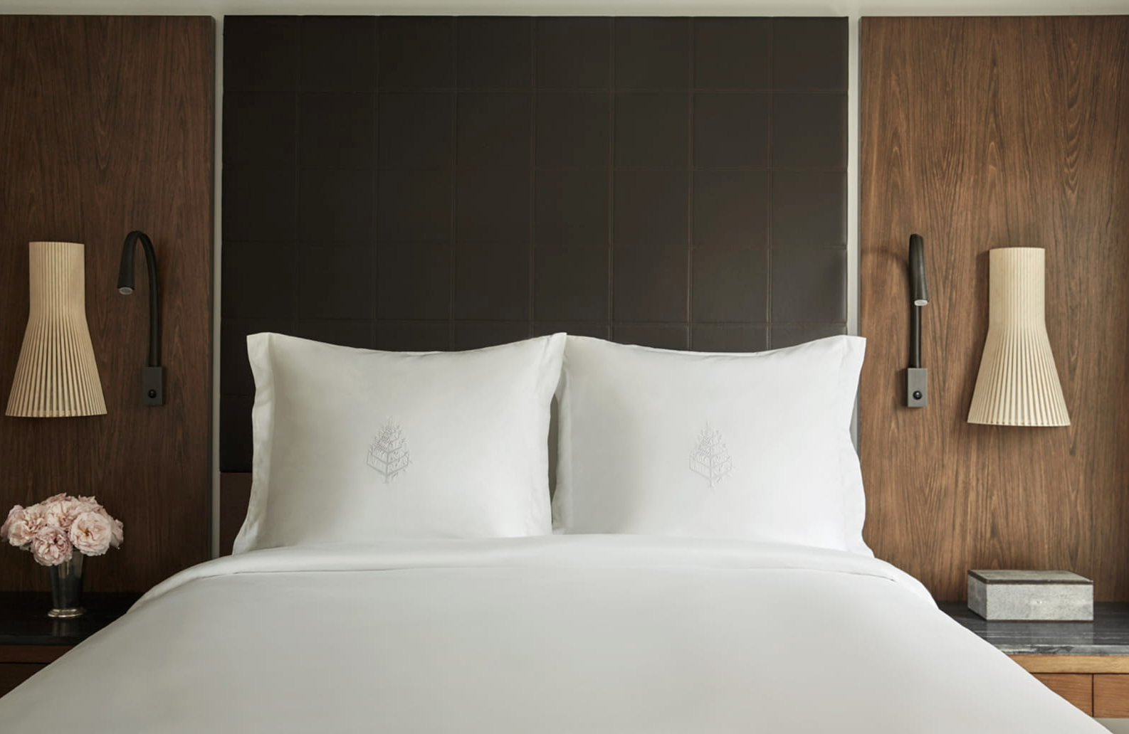 A neatly made bed with white linens and two large pillows with slight embroidery, set against a wooden headboard with two beige wall-mounted lamps on either side and a small nightstand with a vase of pink flowers on the left.
