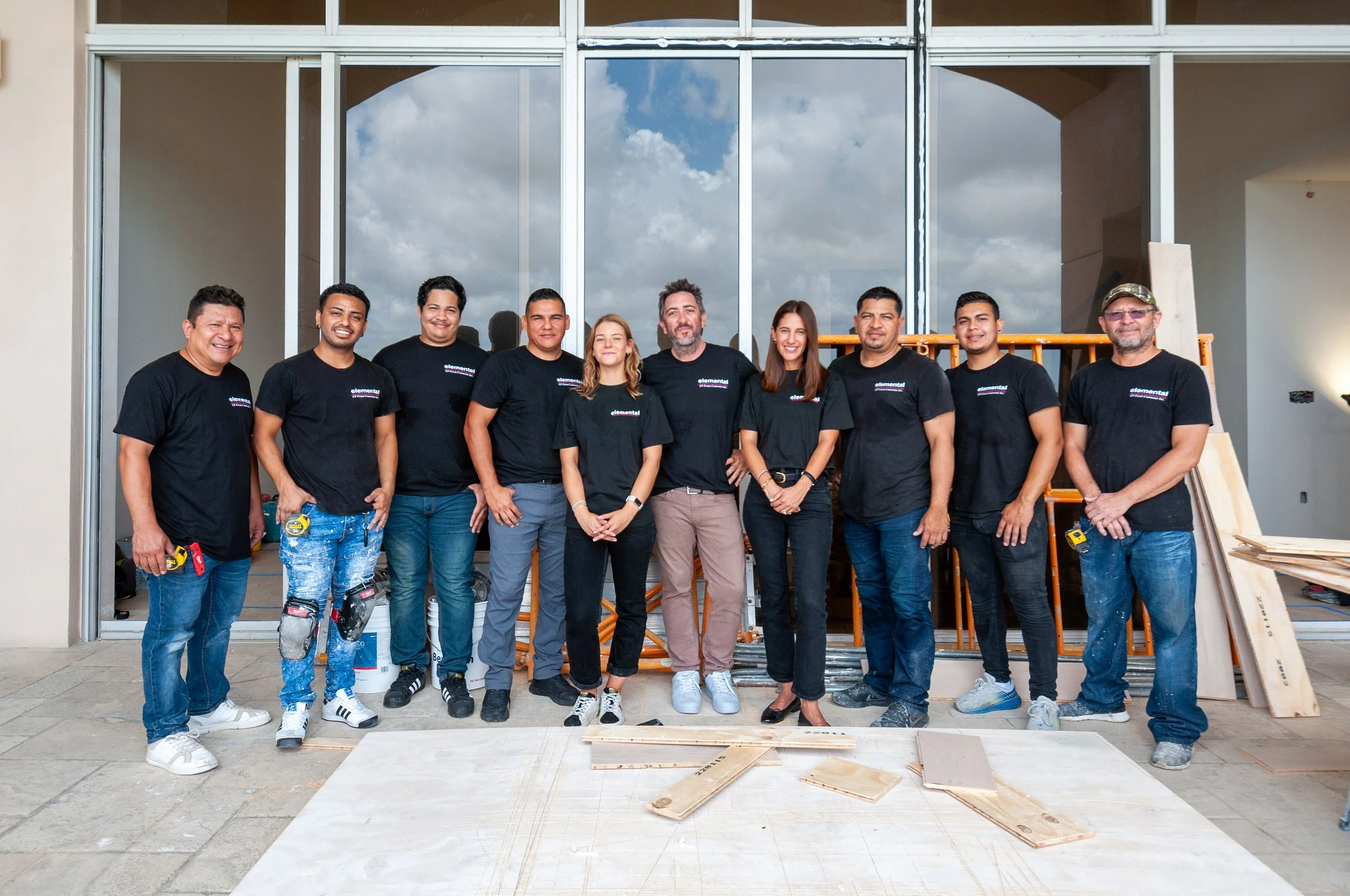 A group of ten diverse people standing in front of a building under construction, smiling at the camera. They are wearing black T-shirts with a logo, some are holding tools, and there is construction equipment and wood scattered on the ground.