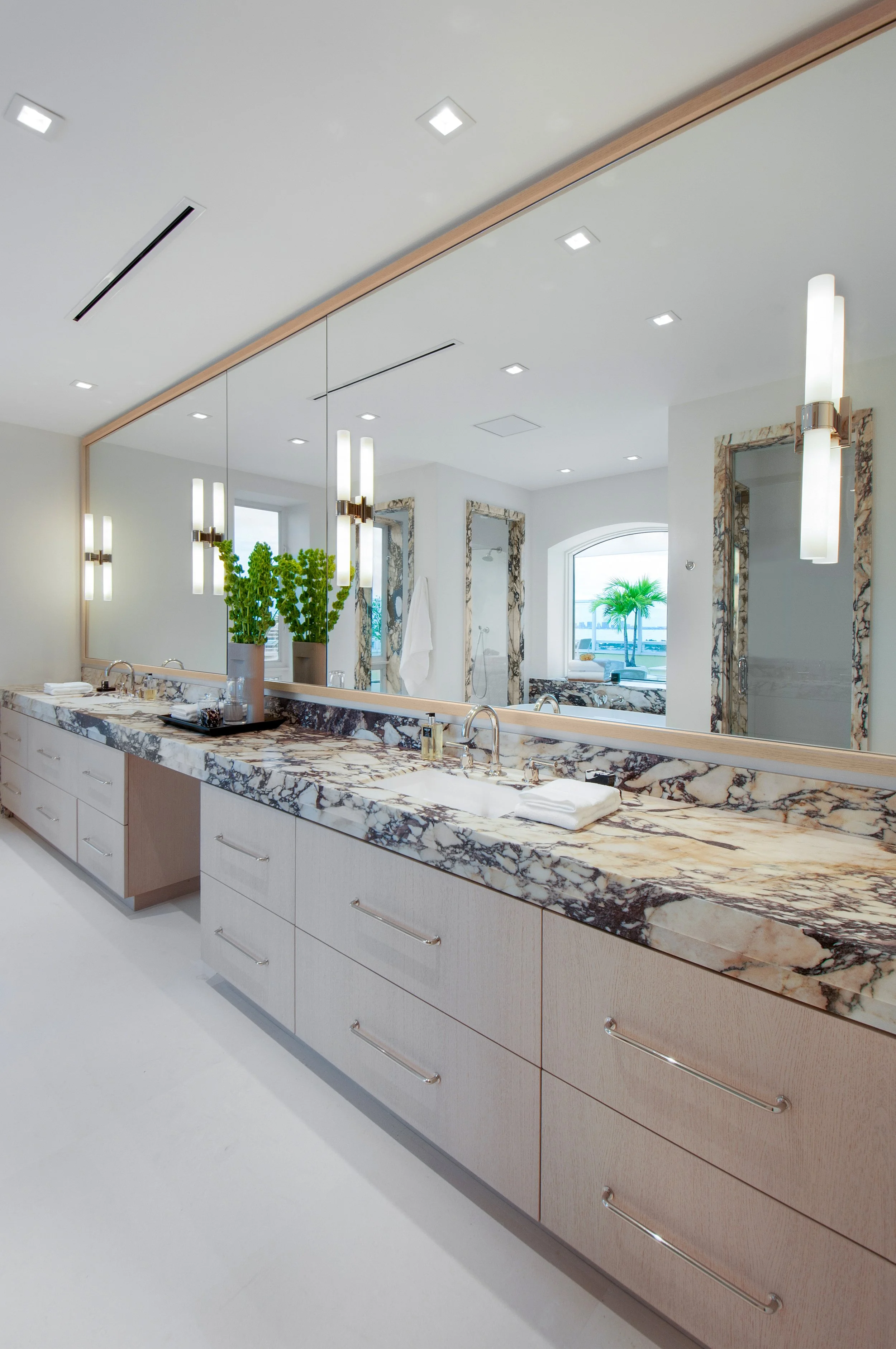 Modern bathroom with white and wood cabinetry, large mirror, marble countertop, and potted plants. The room features recessed lighting and view of a city skyline through a window.
