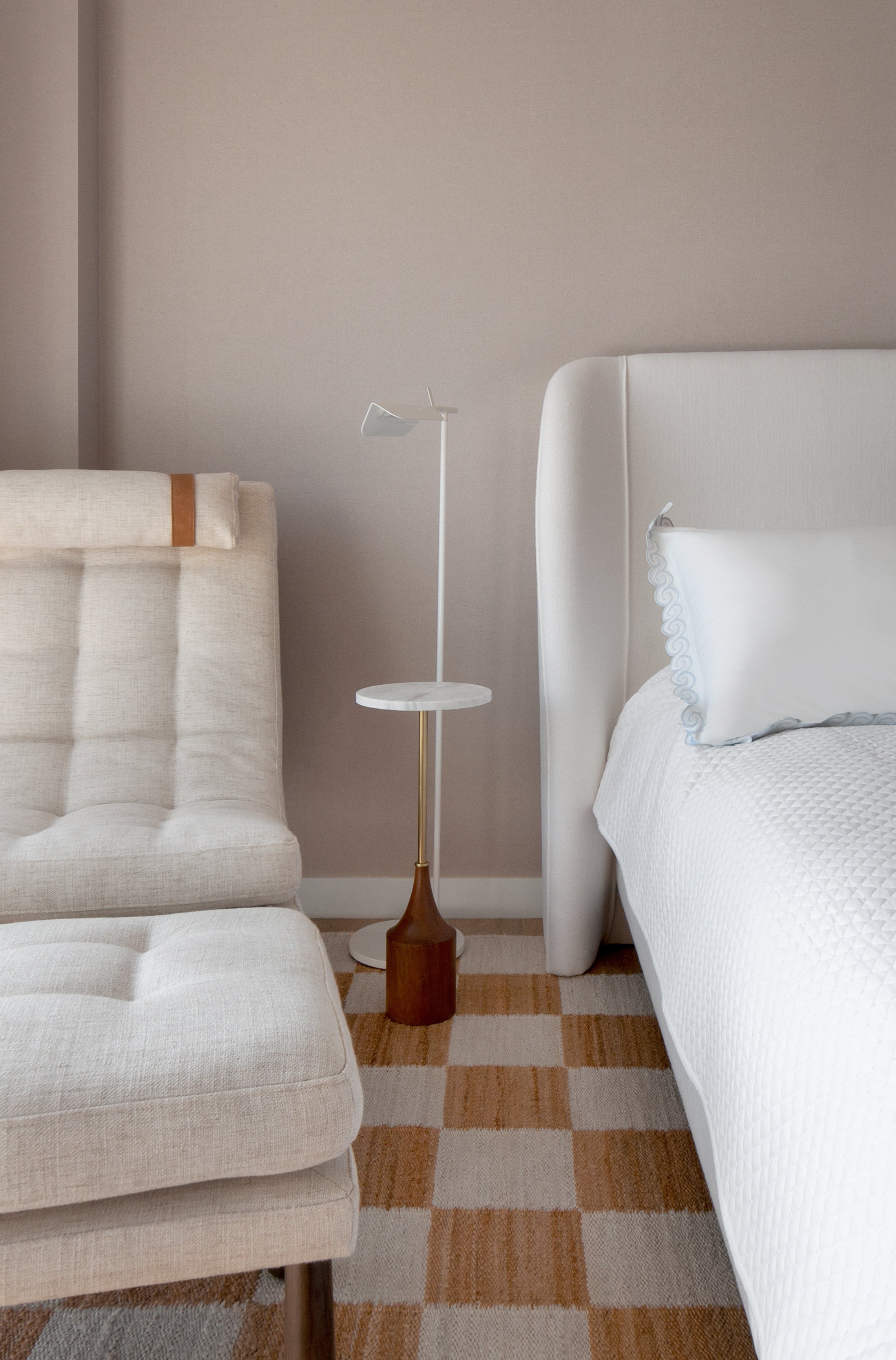 A bedroom scene with a beige tufted armchair on the left, a white bed with a white pillow on the right, and a tall white marble-top lamp with a modern lampshade between them. There is a brown and beige checkered rug on a hardwood floor.
