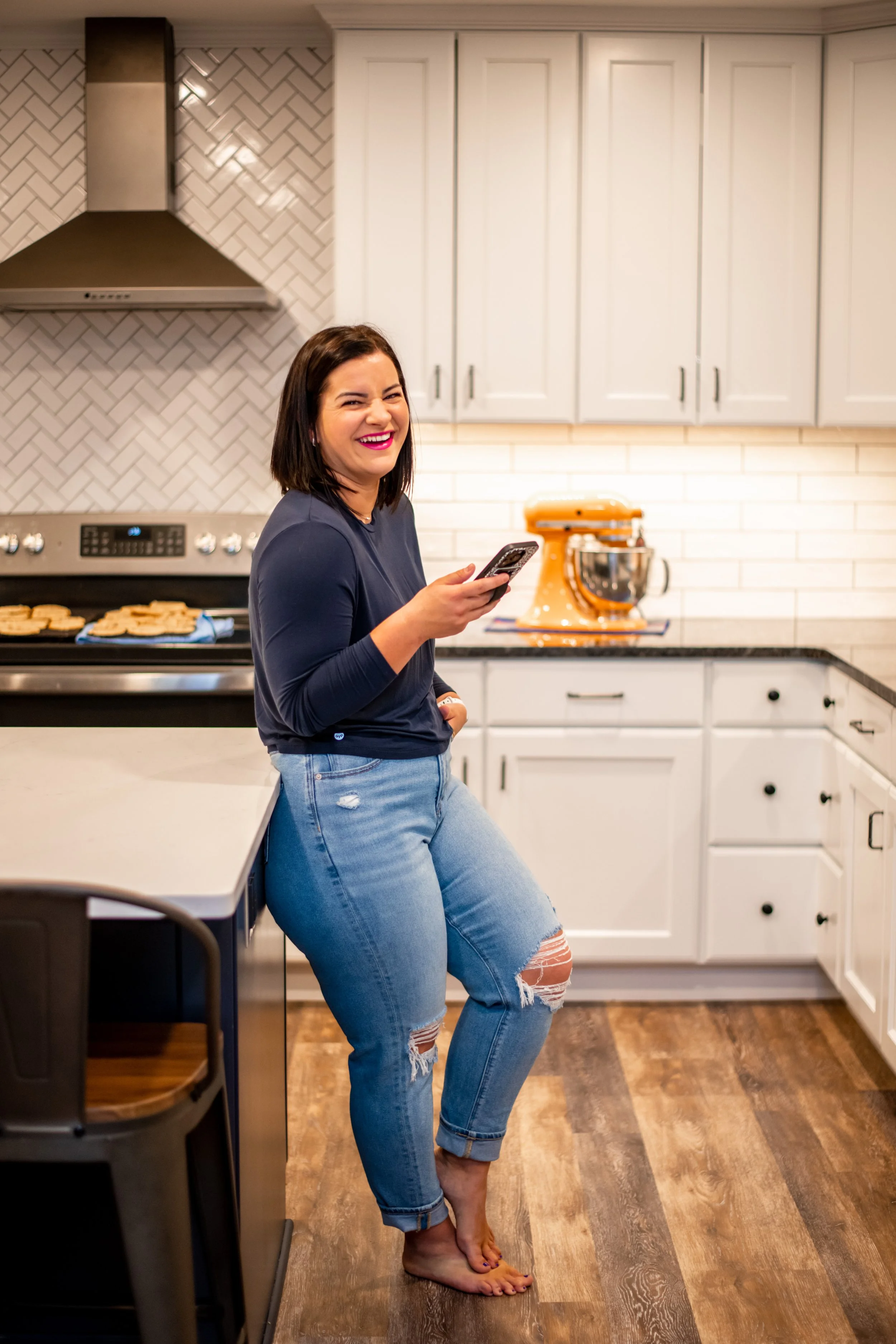 A woman with shoulder-length dark hair, wearing a navy sweater and ripped jeans, smiling and holding a smartphone in a kitchen. She is barefoot, standing on hardwood flooring, with white cabinets, a silver stove, a range hood, and an orange stand mixer in the background.