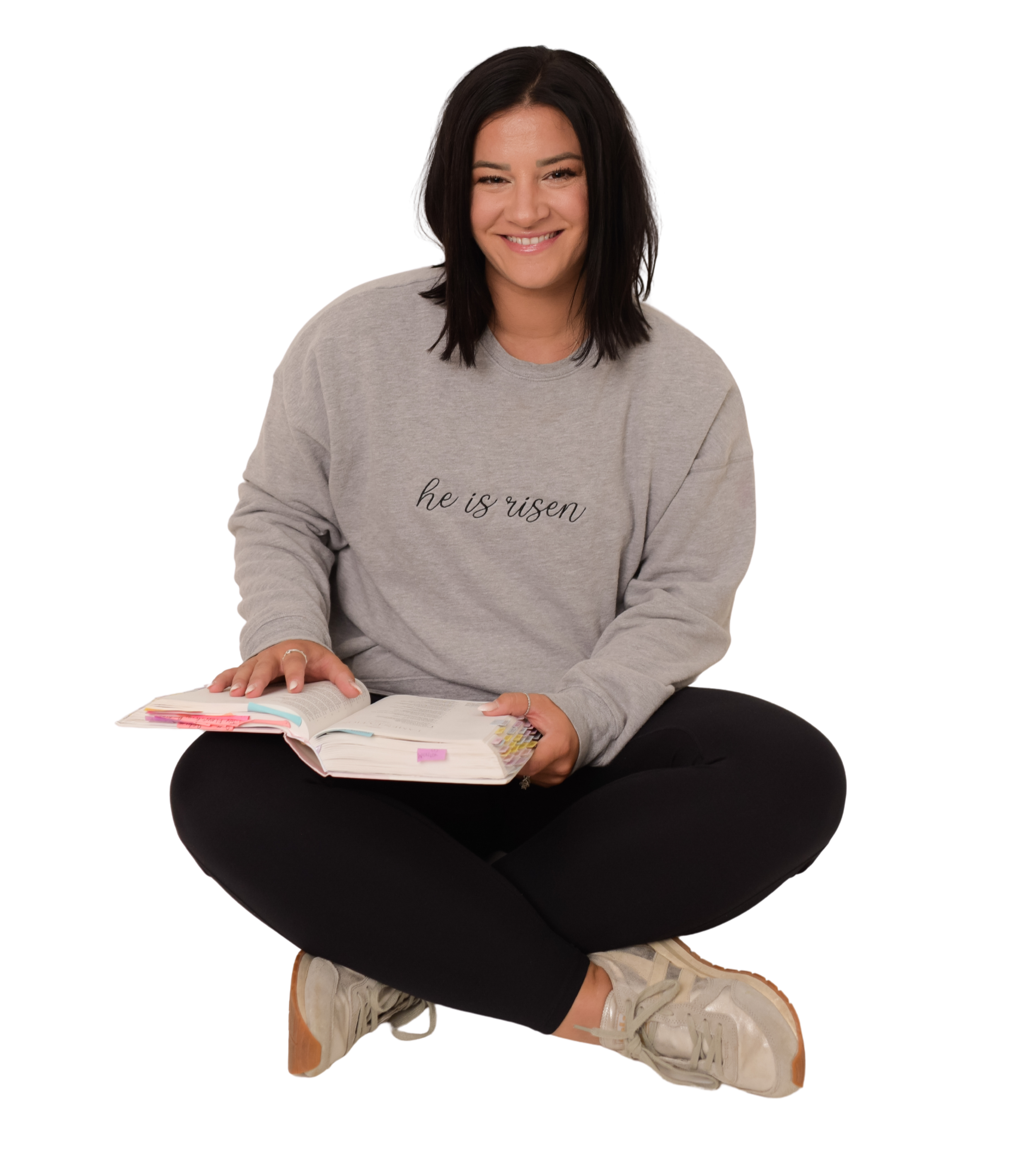 A young woman sitting cross-legged on the floor, smiling at the camera. She is wearing a light gray sweatshirt with the words "he is risen" printed on it, black pants, and beige sneakers. She has dark shoulder-length hair and is flipping through the pages of an open book or notebook.
