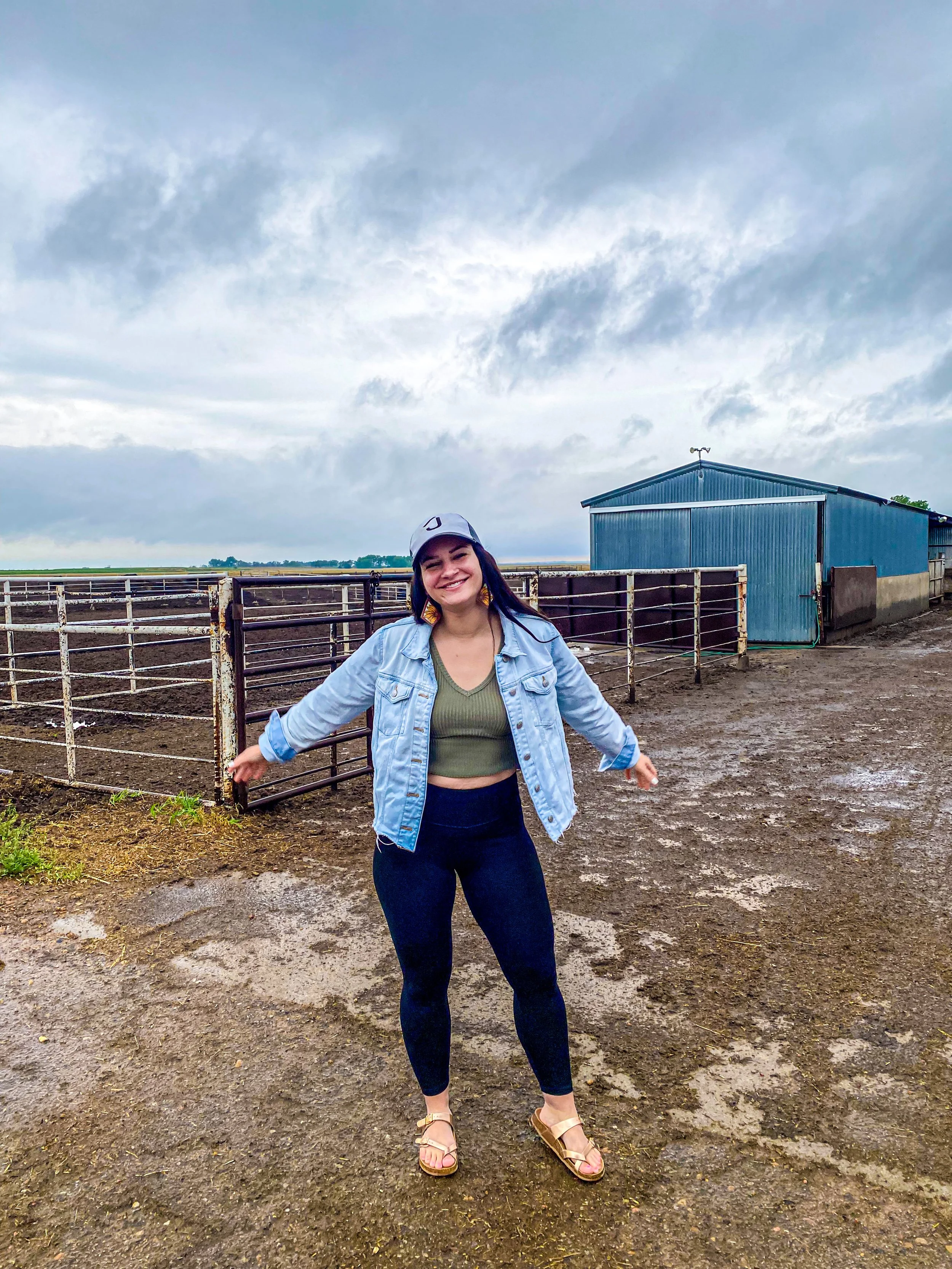 A young woman in a denim jacket, green crop top, and black pants, smiling with her arms outstretched, standing on muddy ground at a farm or ranch with a blue barn and metal fences in the background under a cloudy sky.