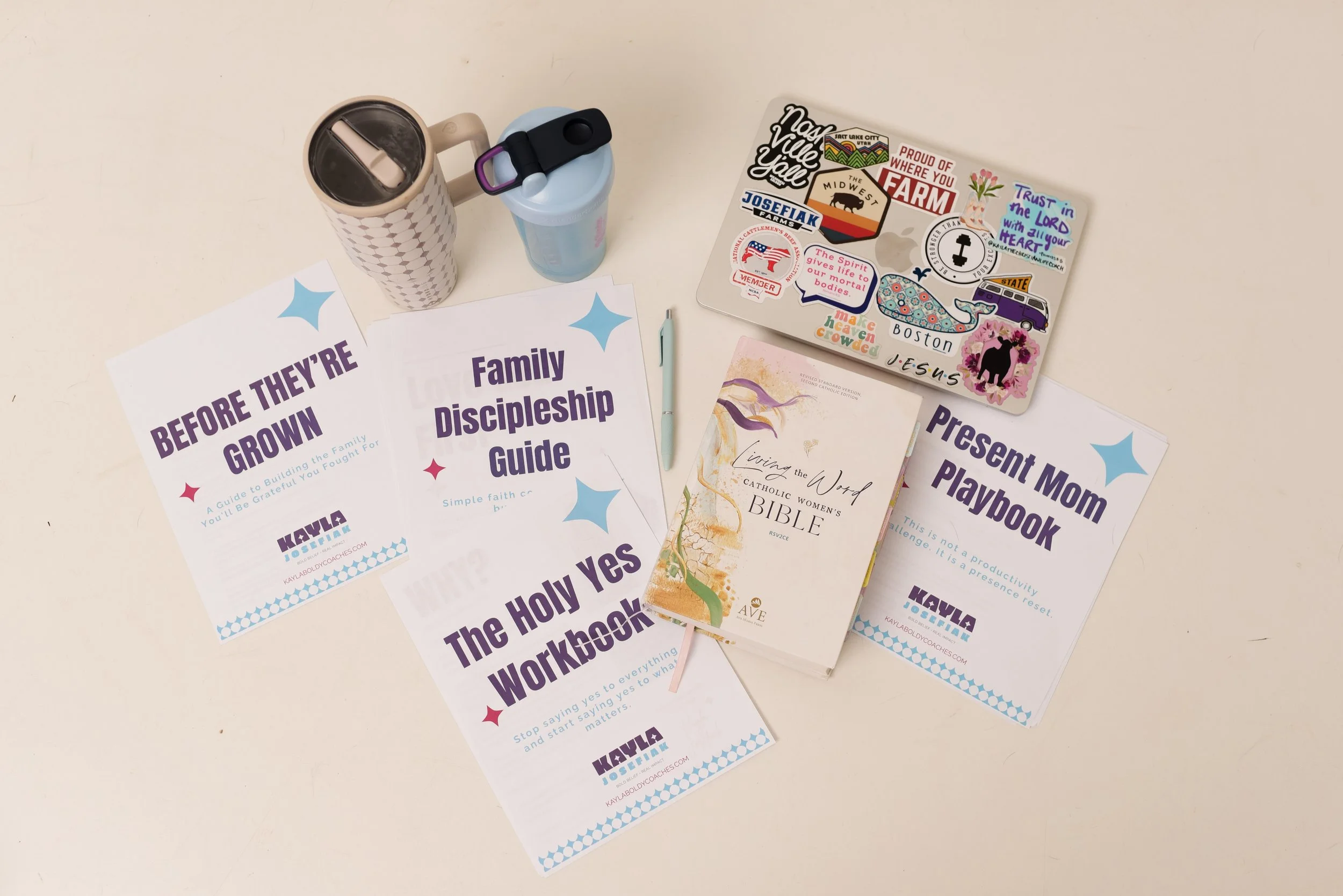 Desk with two water bottles, various printed handouts and booklets about religious and family topics, a laptop covered with stickers, and a pen.