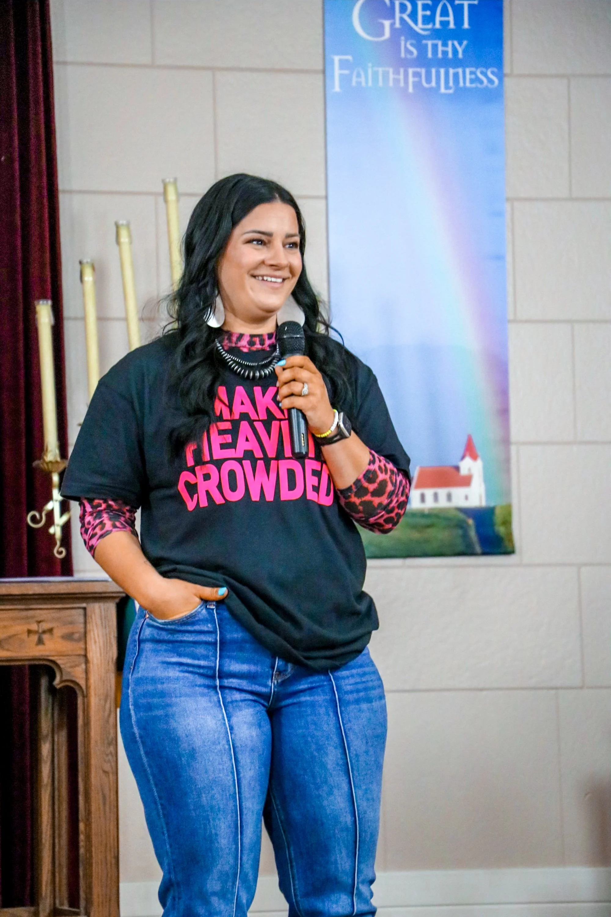 A woman with dark hair, wearing a black T-shirt with pink text and a leopard print long sleeve shirt underneath, standing and smiling while holding a microphone. Behind her is a banner that reads 'GREAT IS THY FAITHFULNESS' and a painting of a church and rainbow.