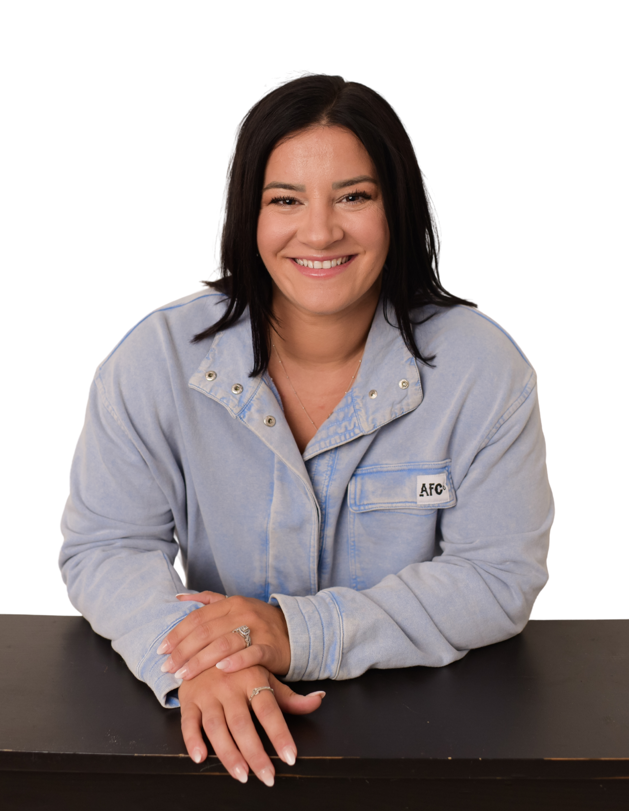 A woman with dark hair smiling, sitting at a black table with her hands clasped, wearing a light denim jacket with a patch reading 'AFC'.