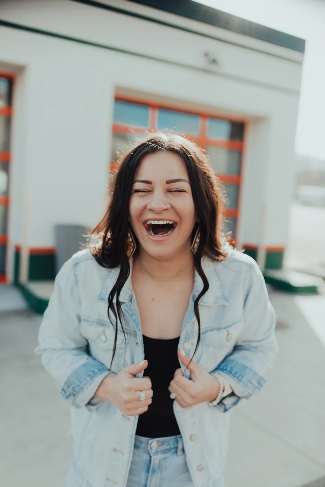 A woman with dark hair laughing outdoors in front of a building with orange-trimmed windows, wearing a denim jacket and black top.