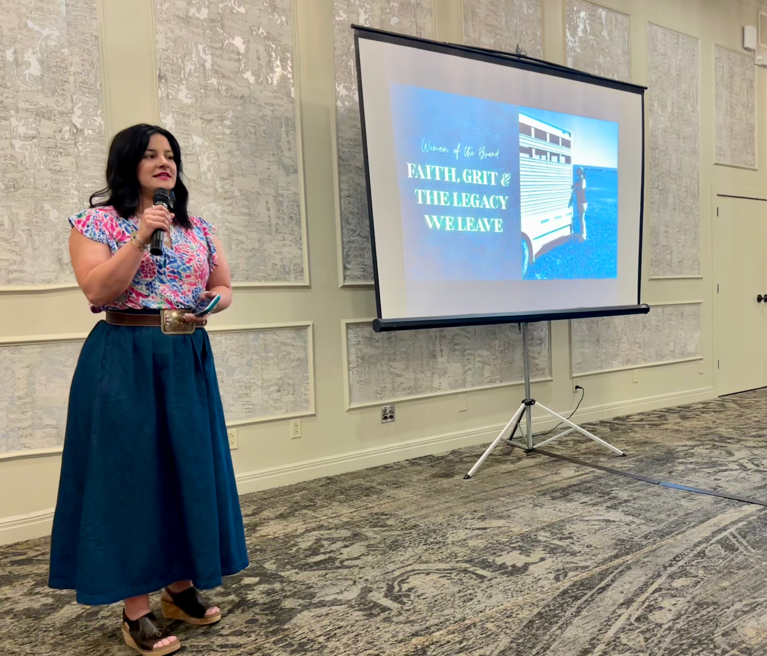 A woman standing in front of a presentation screen, holding a microphone, with a sign on the screen that reads, "Women of the Brand: Faith, Grit & the Legacy We Leave." She is wearing a colorful top and a long, dark skirt.