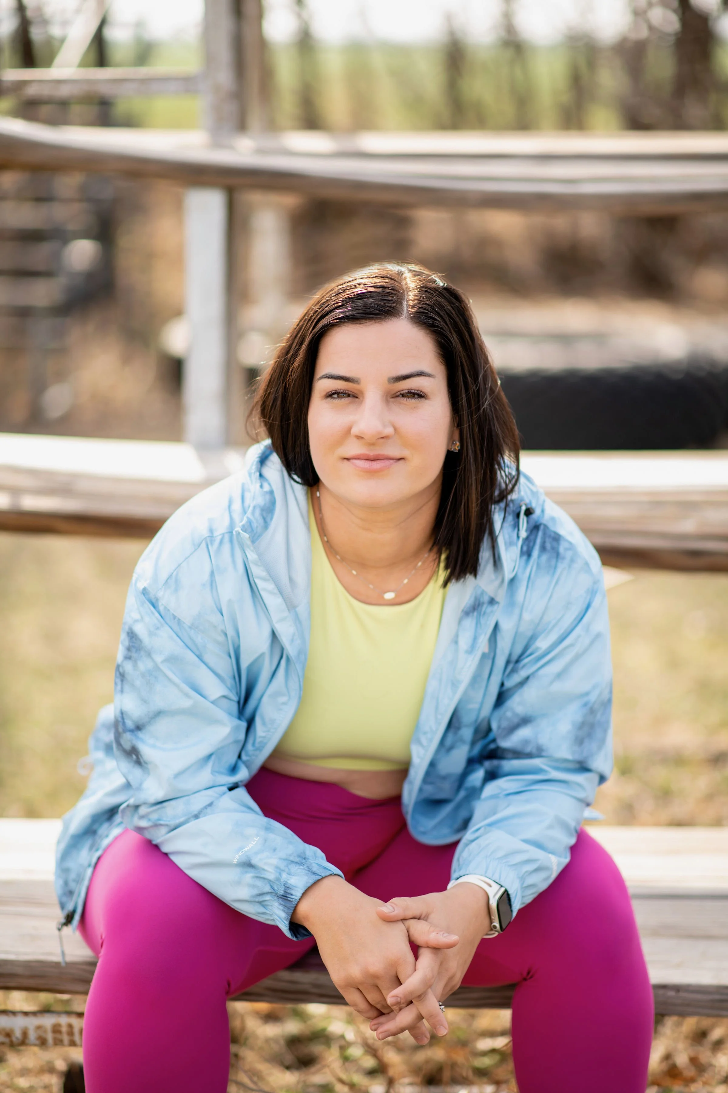 A woman with dark hair and light skin, wearing a yellow top, pink leggings, and a light blue jacket, sitting on a wooden park bench outdoors. She is smiling slightly and looking at the camera with trees and a blurred background.