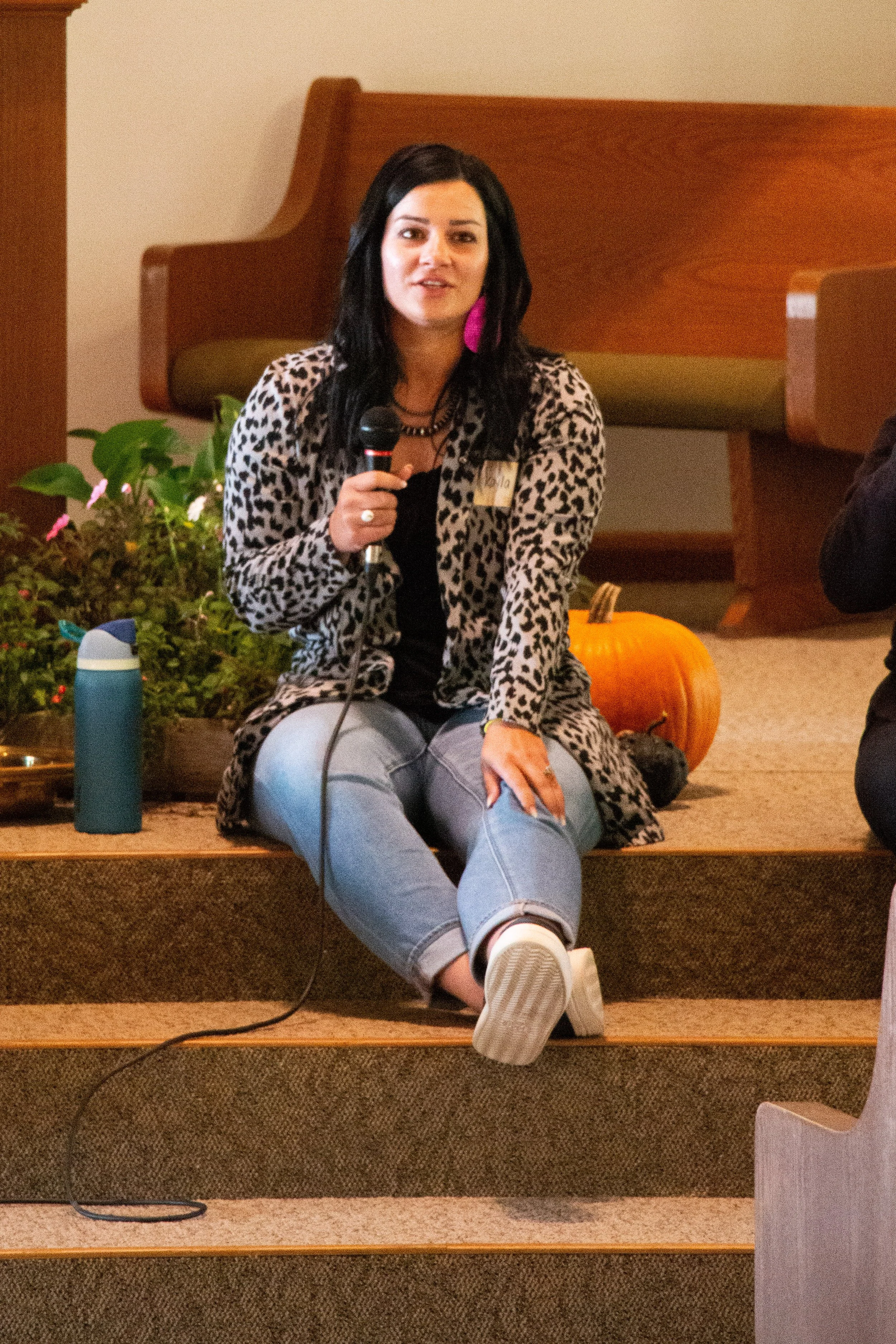 A woman with black hair wearing a leopard print coat and white pants is sitting on a stage with a microphone in her hand, in front of some pumpkins and plants.