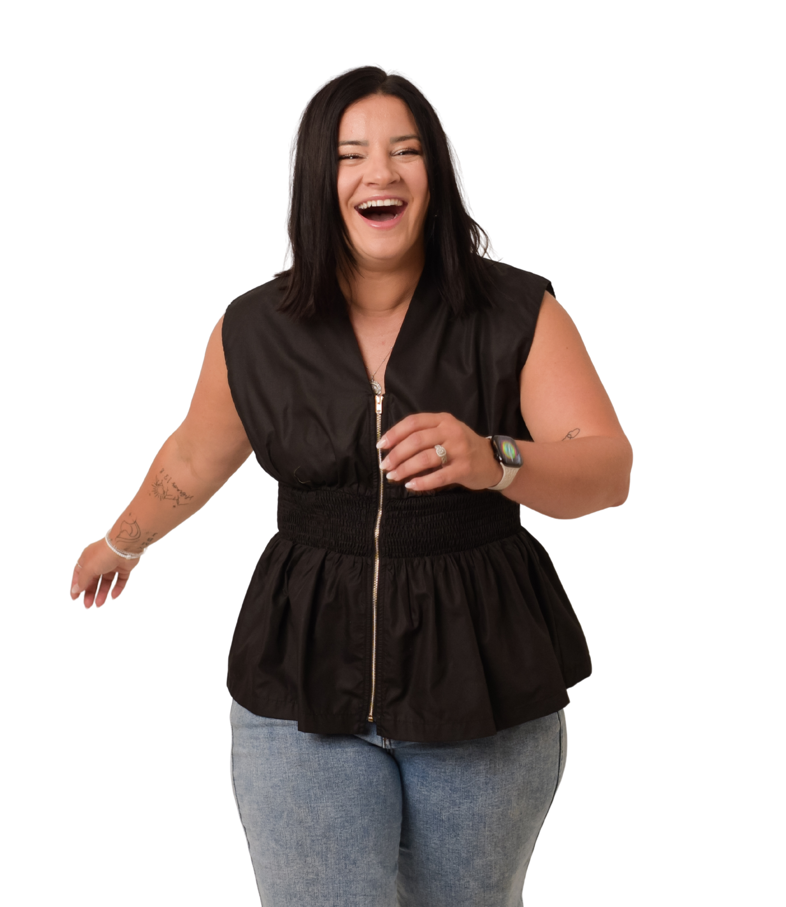 A woman with dark hair, wearing a black sleeveless zip-up top, blue jeans, and a smartwatch, laughing and leaning forward with tattoos on her left arm.
