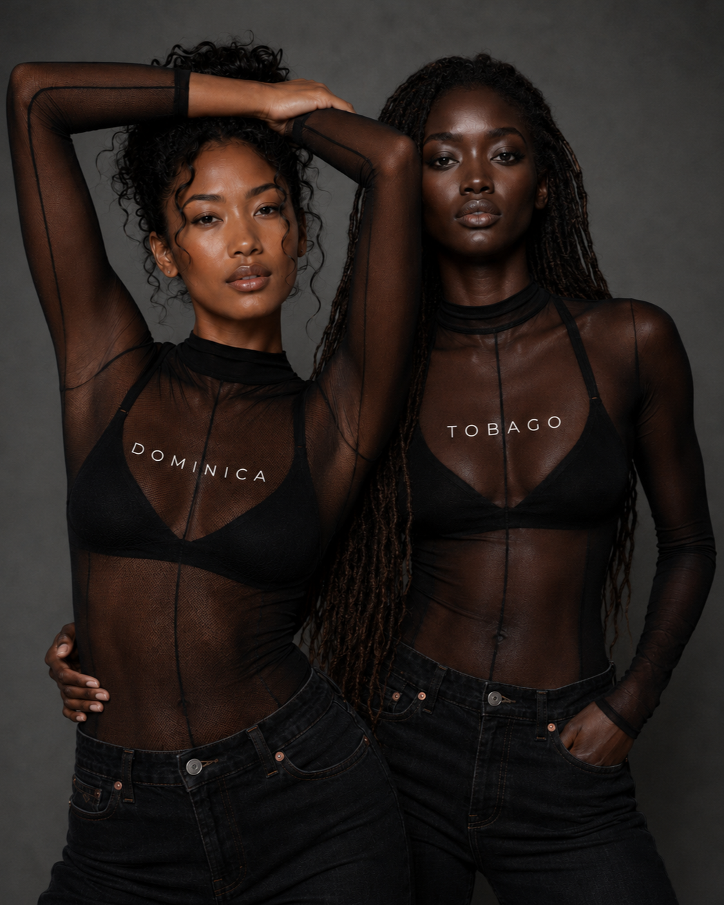Two women with dark skin and curly and braided hair wearing sheer black tops with country names 'Dominica' and 'Tobago' and black jeans, standing against a gray background.