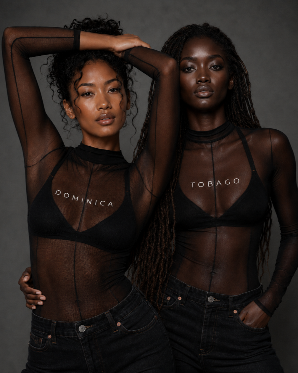 Two women with dark skin and curly and braided hair wearing sheer black tops with country names 'Dominica' and 'Tobago' and black jeans, standing against a gray background.