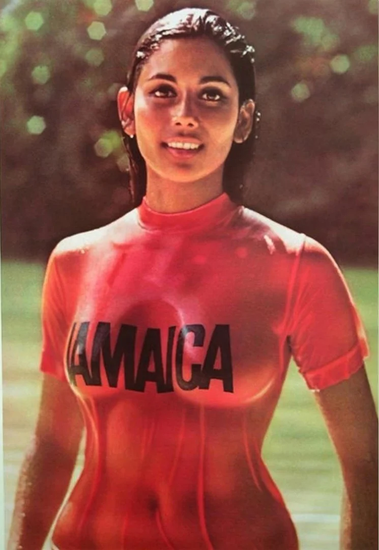 Young woman with wet hair standing outdoors, wearing a red sports shirt that says "JAMAICA" on it, smiling with a blurred green background.
