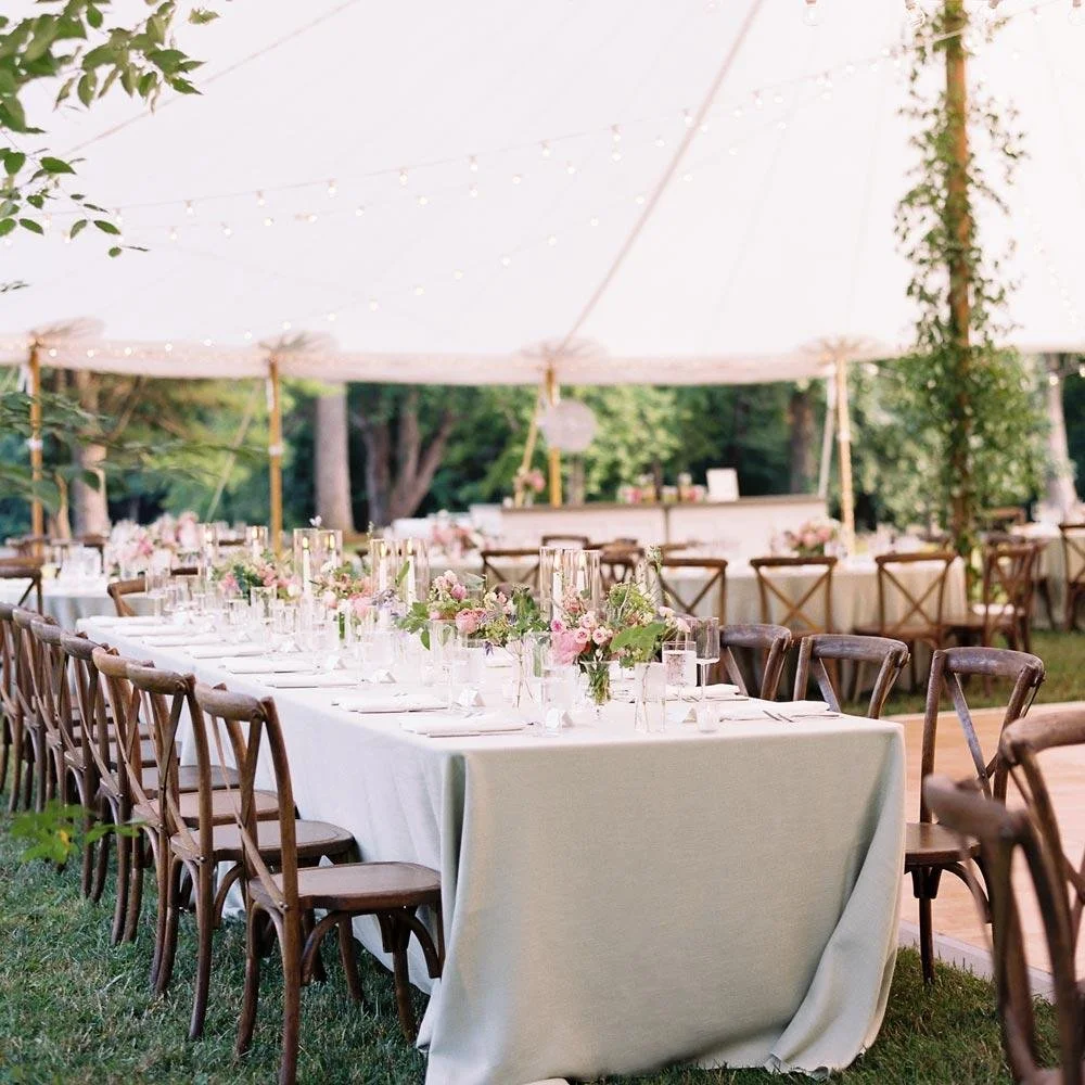Outdoor party setup with a long rectangular table covered with a white tablecloth, decorated with pink and white flowers in glass vases, set with glasses and plates, under a large white tent with string lights, surrounded by trees and wooden chairs.