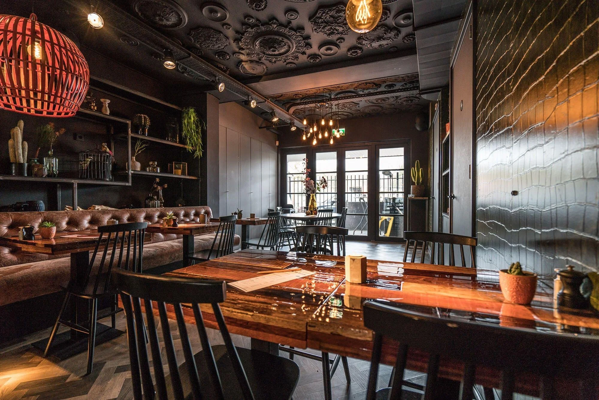Interior of a modern restaurant with wooden tables, black chairs, a tufted bench, and decorative plants on shelves. Large windows let in natural light, with an ornate black ceiling featuring decorative molding and hanging Edison bulb lights. The right wall has a textured, shiny surface.