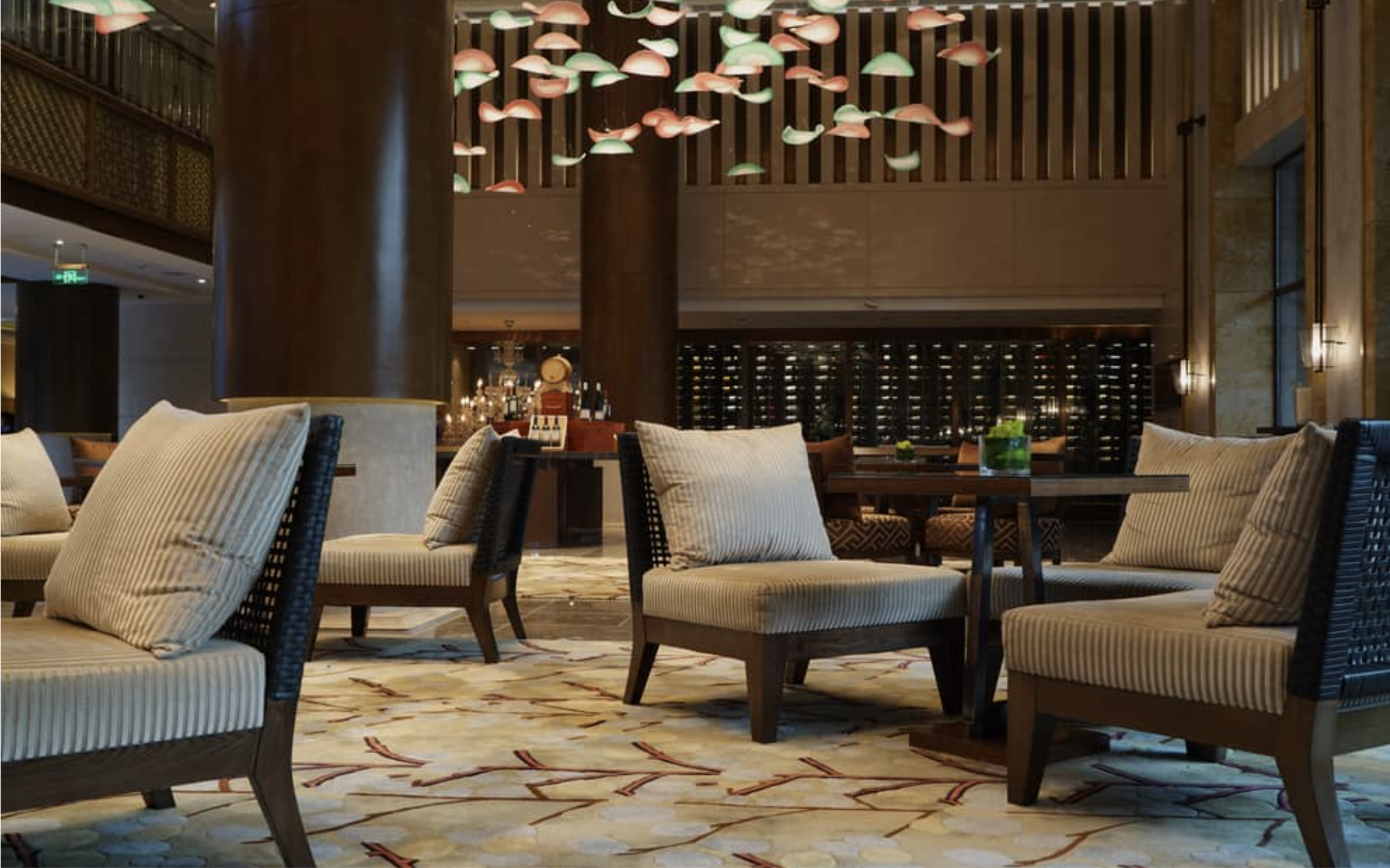 Interior of a restaurant or hotel lounge with beige upholstered chairs, a large decorative rug, and a modern hanging light fixture with pink and green glass shapes.