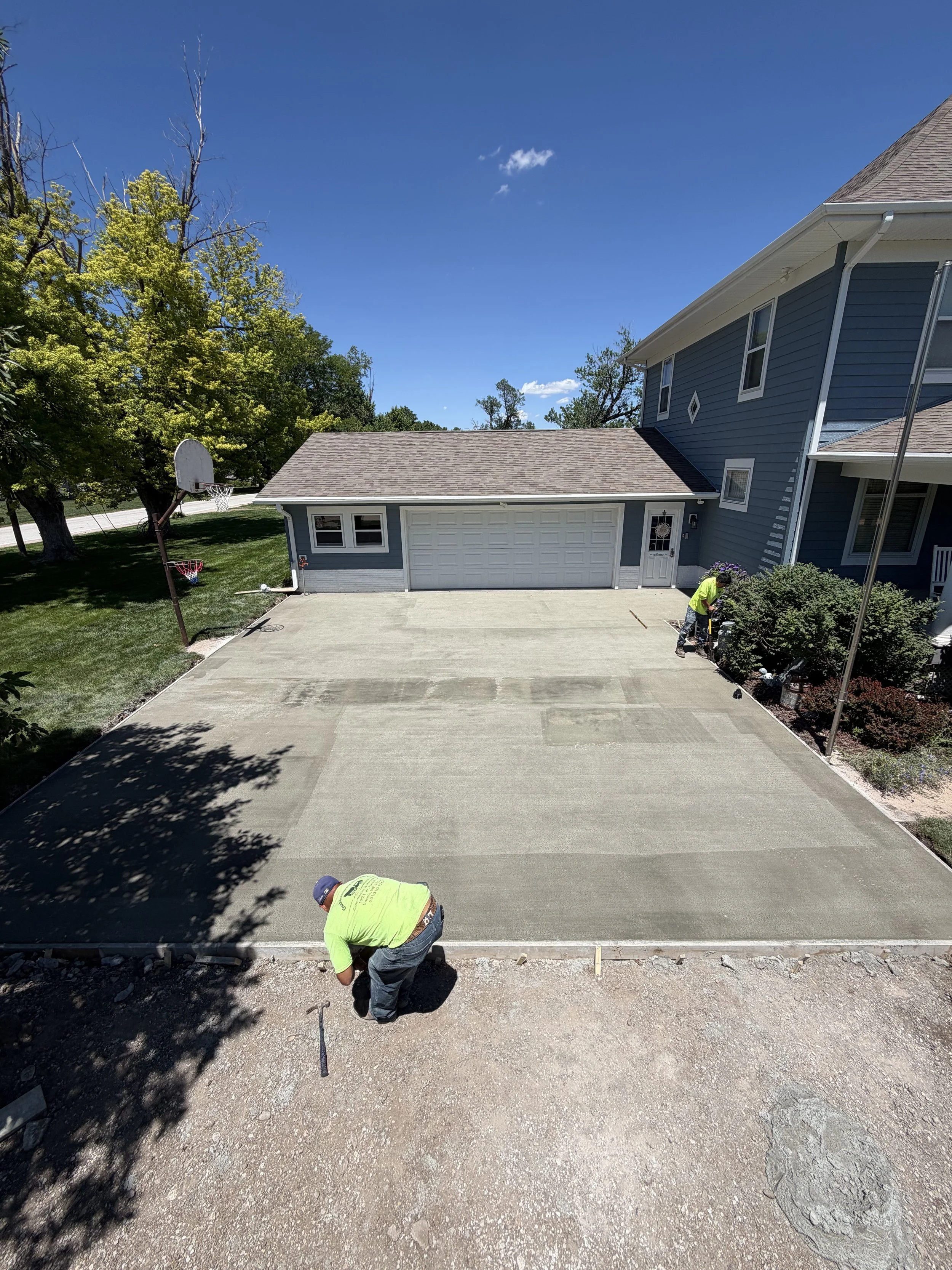 Workers installing a new concrete driveway in front of a blue house with a garage, surrounded by green trees on a sunny day.