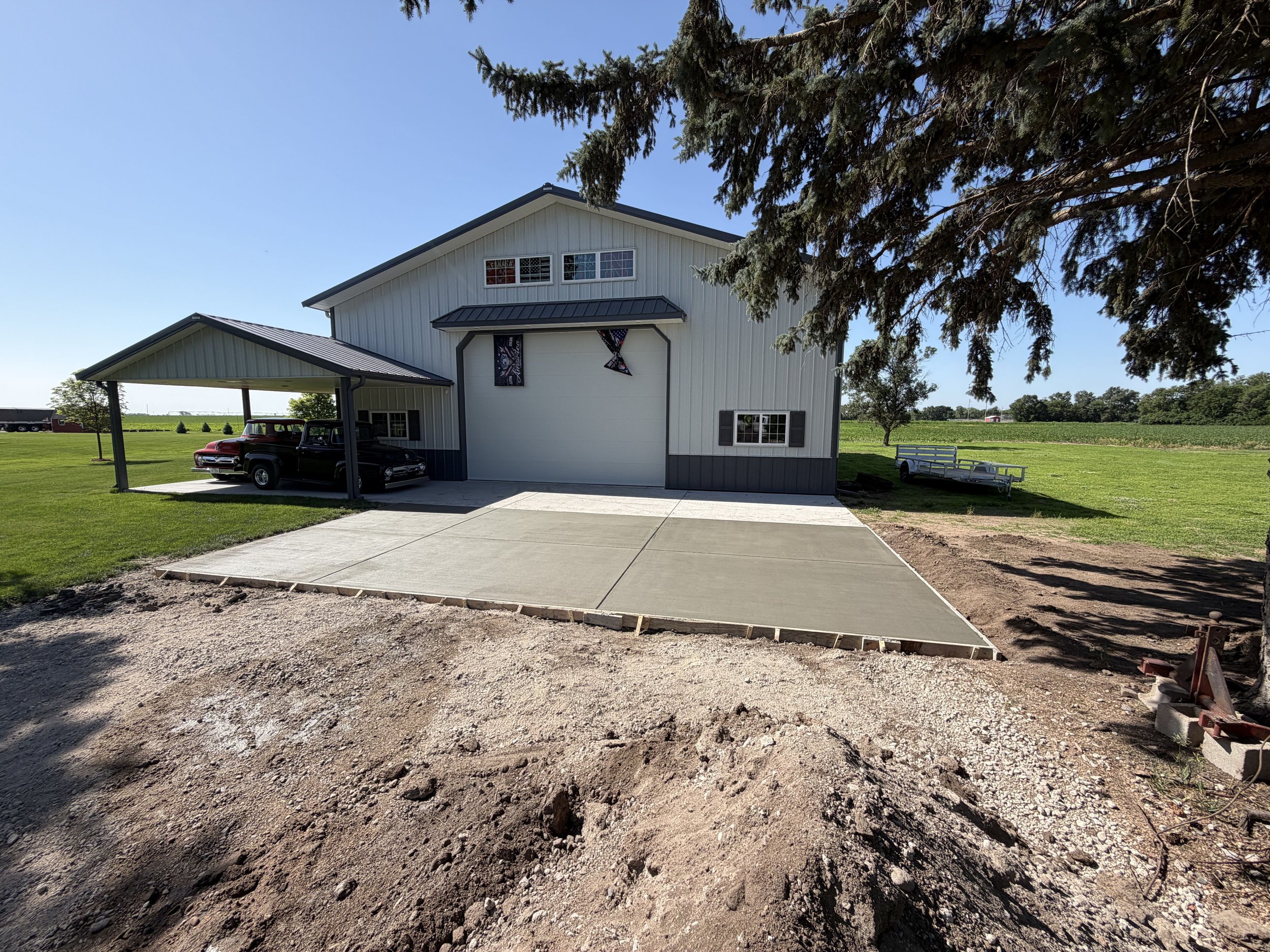 Residential property with white house, cars in carport, landscaped lawn, trees, and a trailer, under clear blue sky.