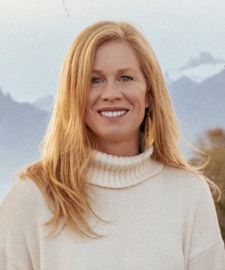 A woman with red hair, smiling, wearing a cream-colored turtleneck sweater, standing outdoors with mountains in the background.