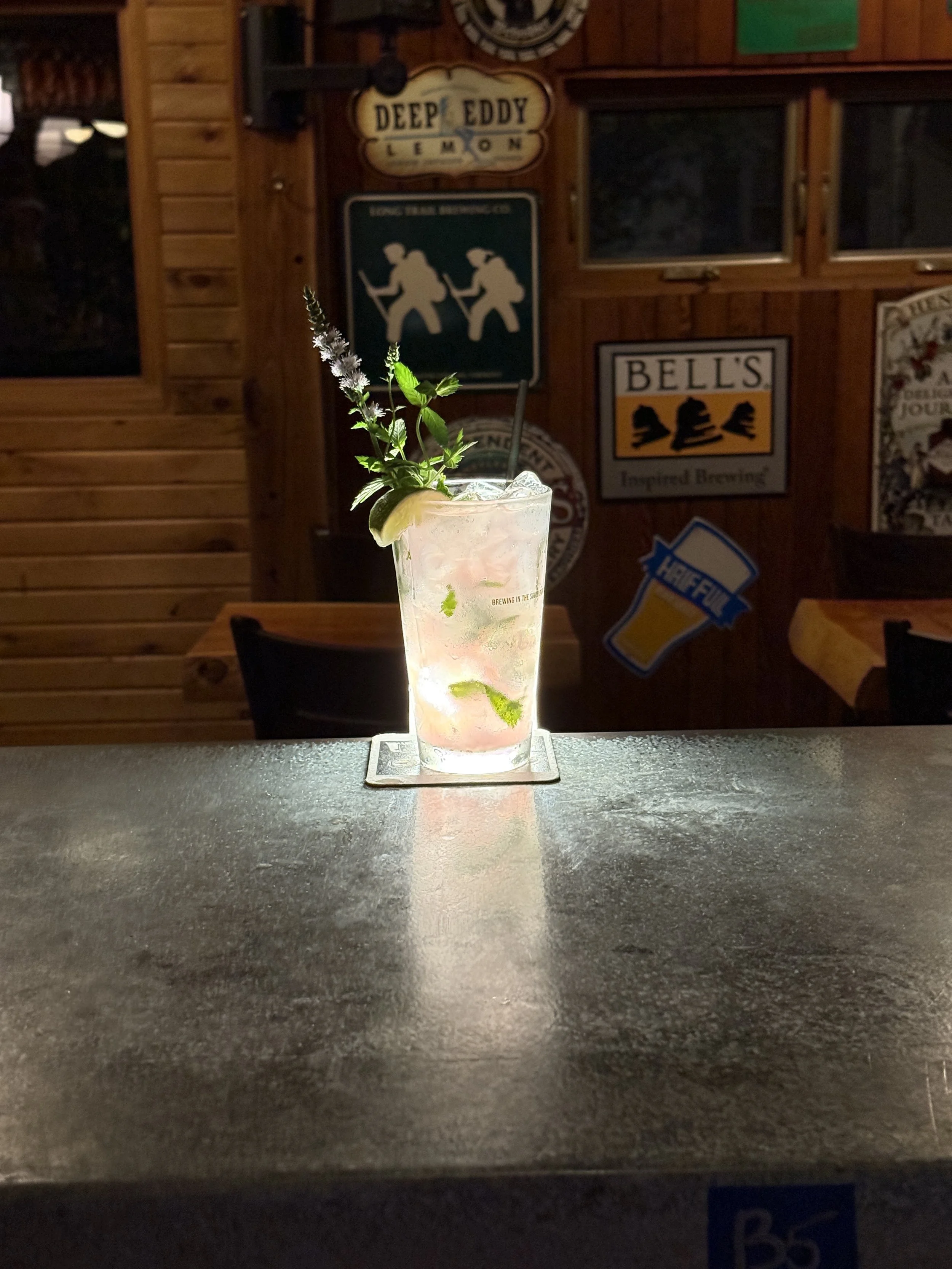 A cocktail garnished with a sprig of herbs, lime wedge, and a straw, on a coaster, in a dimly lit bar or pub with wood-paneled walls decorated with vintage signs and posters.