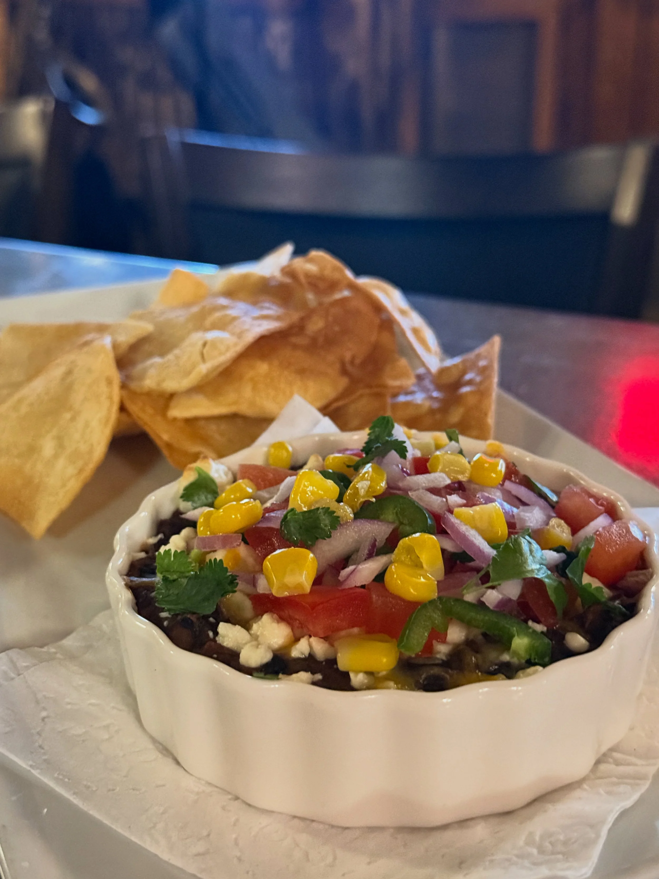 A bowl of colorful Mexican-style salad topped with cilantro, with tortilla chips on the side, on a white surface in a restaurant setting.