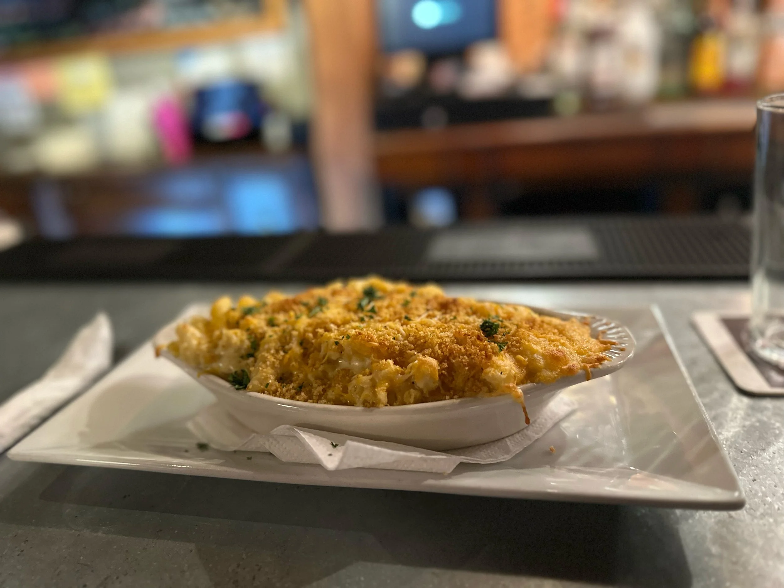 A baked dish of macaroni and cheese with a crispy breadcrumb topping served in a small white oval dish on a tray, with a glass of water to the right, blurred background of what appears to be a restaurant or cafe setting.