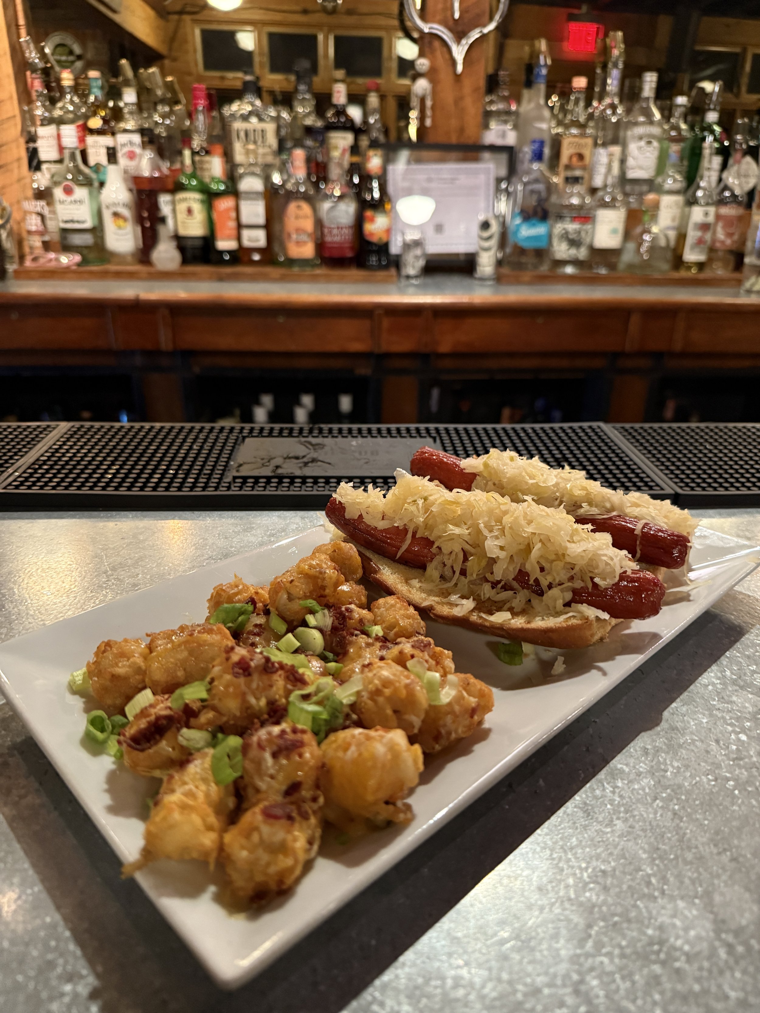 A plate of food with fried chicken pieces garnished with chopped green onions and a hot dog topped with sauerkraut, served on a white rectangular dish in front of a bar counter filled with various liquor bottles in a rustic wooden bar setting.