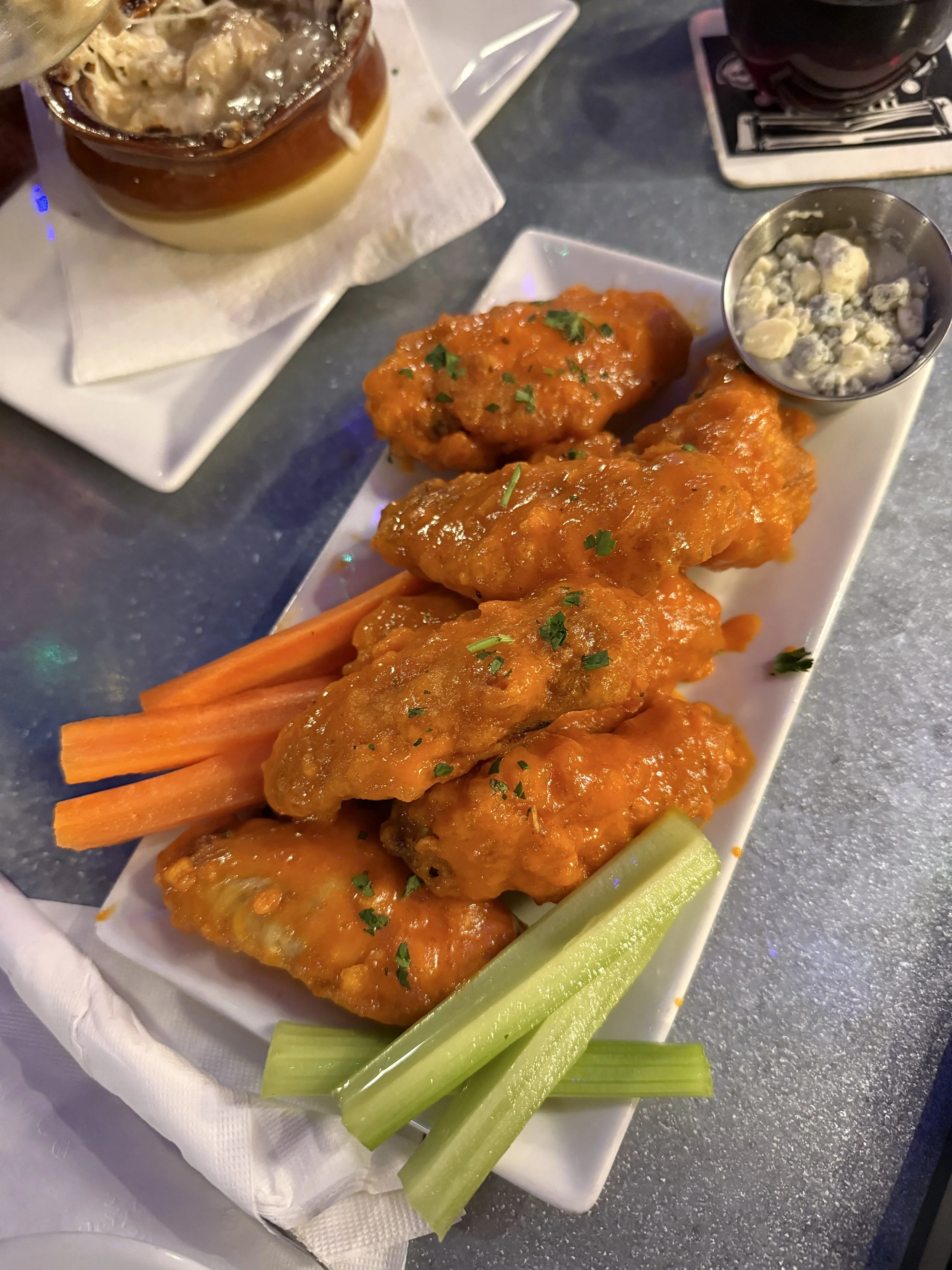 A white rectangular plate with buffalo chicken wings garnished with chopped parsley, celery sticks, and a side of blue cheese crumbles; in the background, a glass jar with a layered dessert and a dark beverage on a coaster.