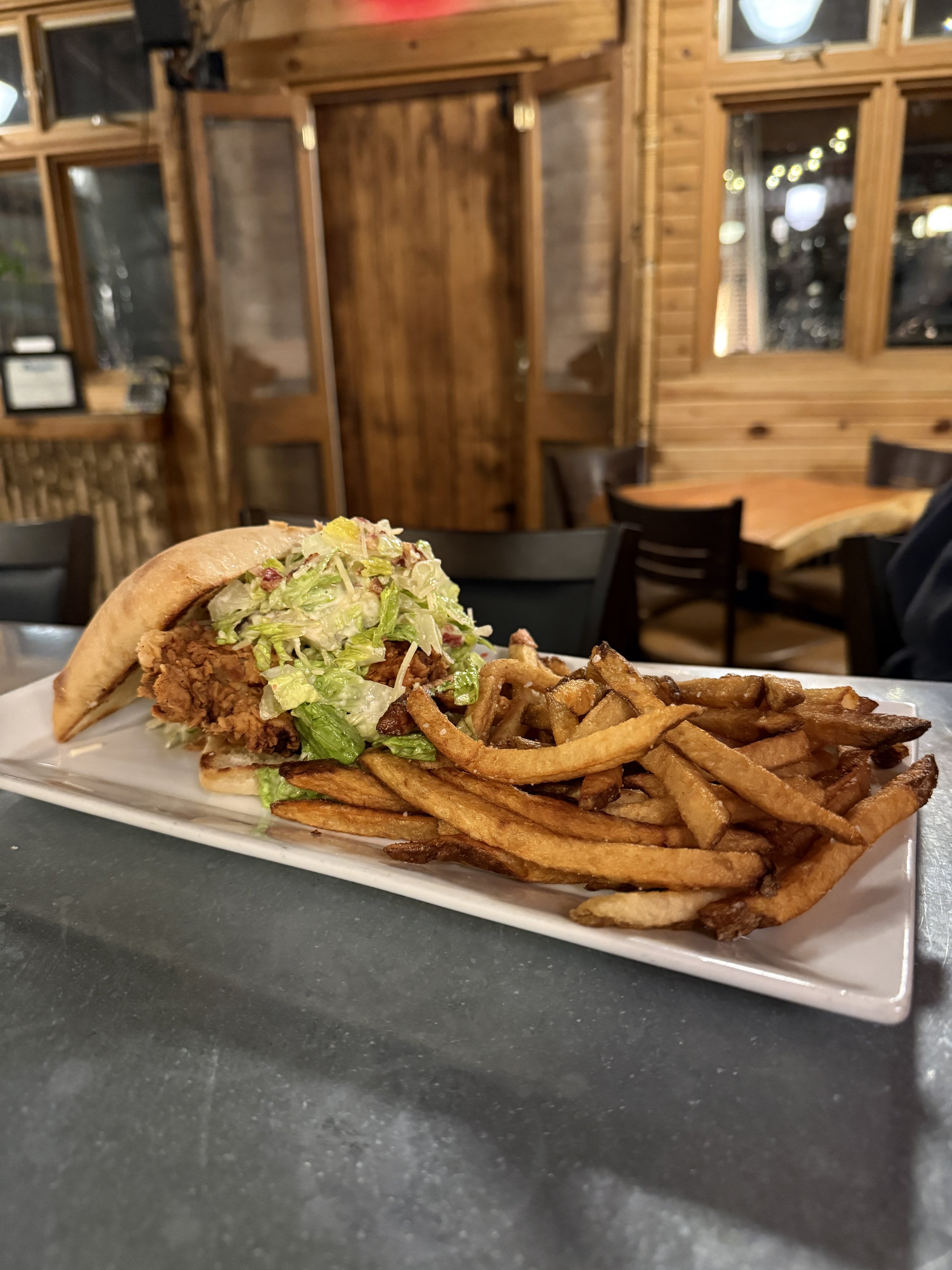 A plate with a sandwich containing fried chicken, shredded lettuce, and tomato, alongside a serving of French fries, on a dark table in a restaurant with wooden walls and windows.