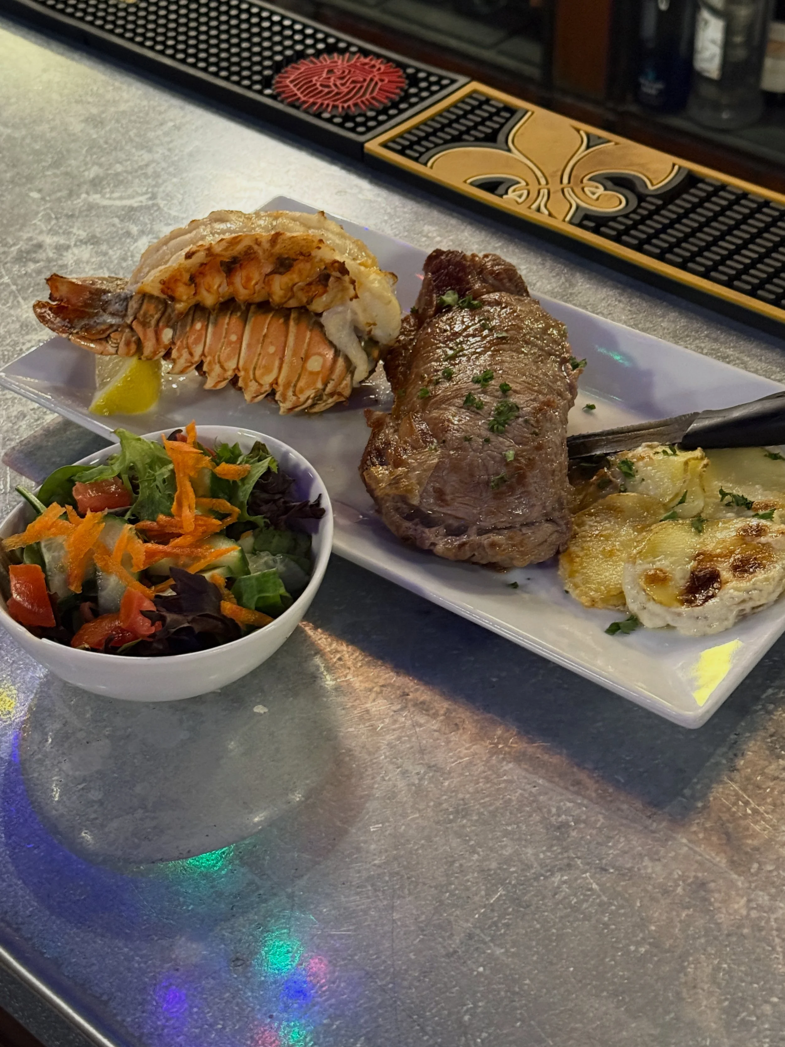 A white rectangular plate with grilled lobster tail and a beef short rib on a gray countertop. There is a small bowl of mixed salad with shredded carrots and greens next to the plate. In the background, a bar mat with a red logo is visible.