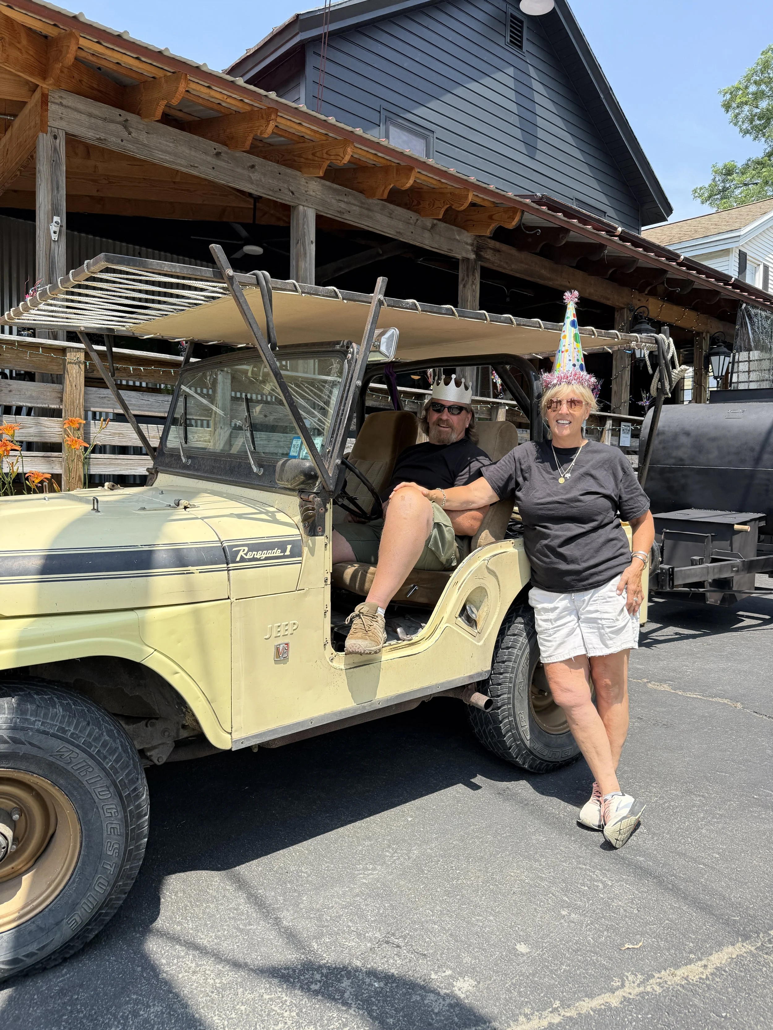 Two people celebrating on a vintage yellow Jeep Wrangler, wearing birthday hats, with a decorated porch and house in the background.