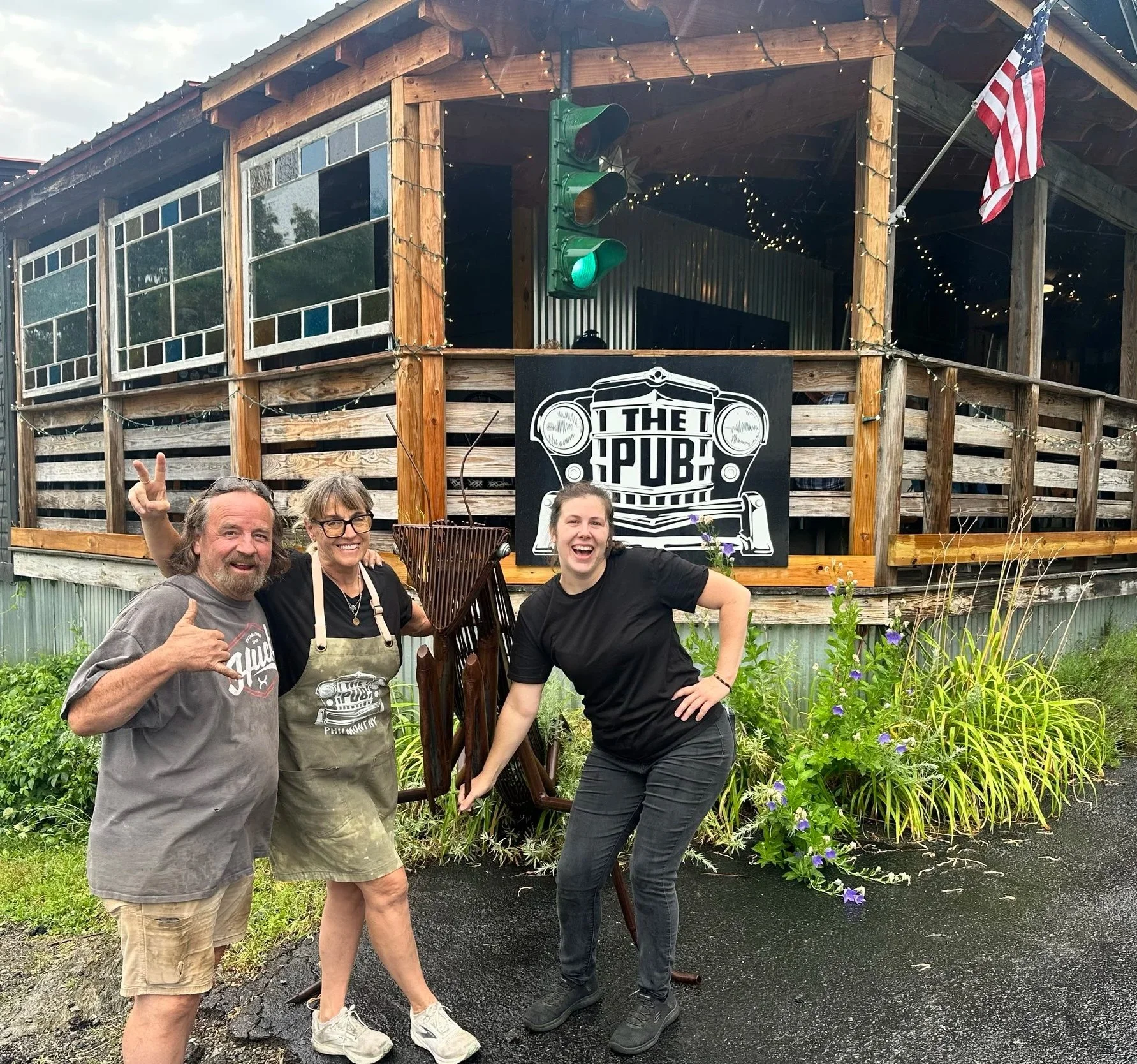 Three smiling people standing outside in front of a wooden building with a sign that says 'The Pub.' The building has string lights, a green traffic light, and an American flag. The group is holding a vintage-style basket and posing happily, with plants and flowers in the foreground.