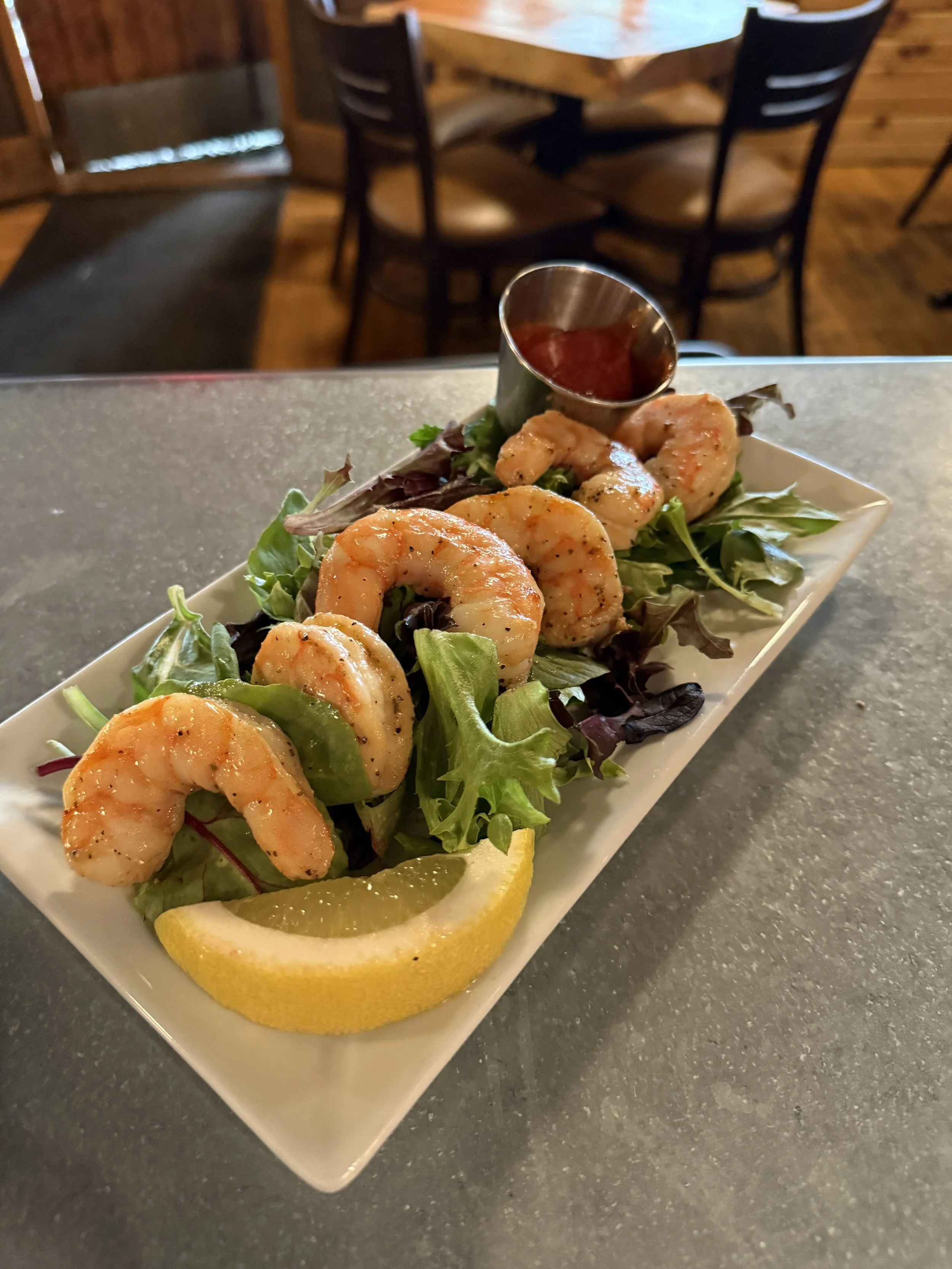 Seafood salad with shrimp, mixed greens, a lemon wedge, and a small cup of cocktail sauce, served on a rectangular white plate.