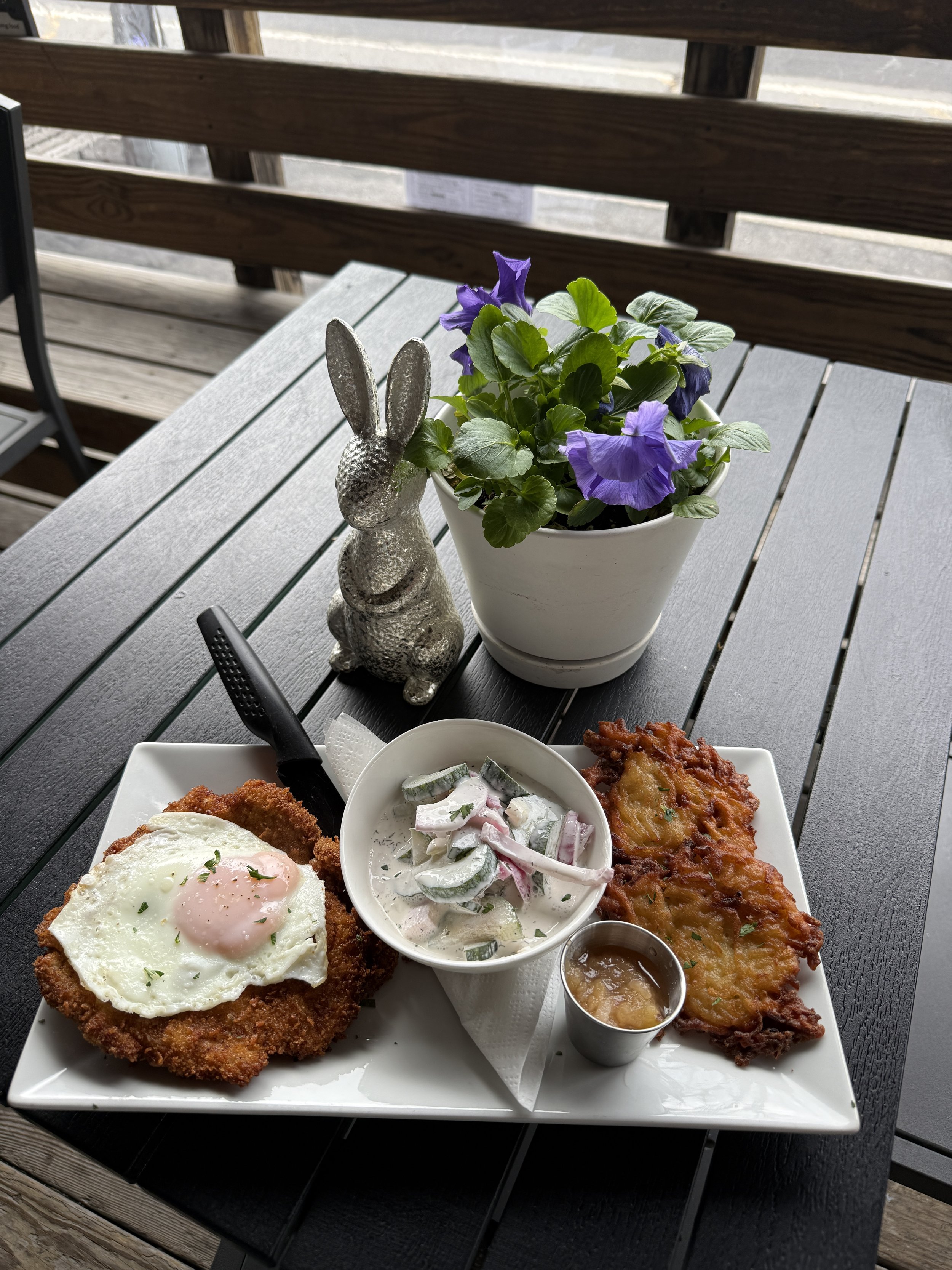 Plate of fried chicken with a fried egg on top, a side of cucumber salad, and a serving of baked or fried dish, with a small cup of sauce, on a gray wooden table outdoors.