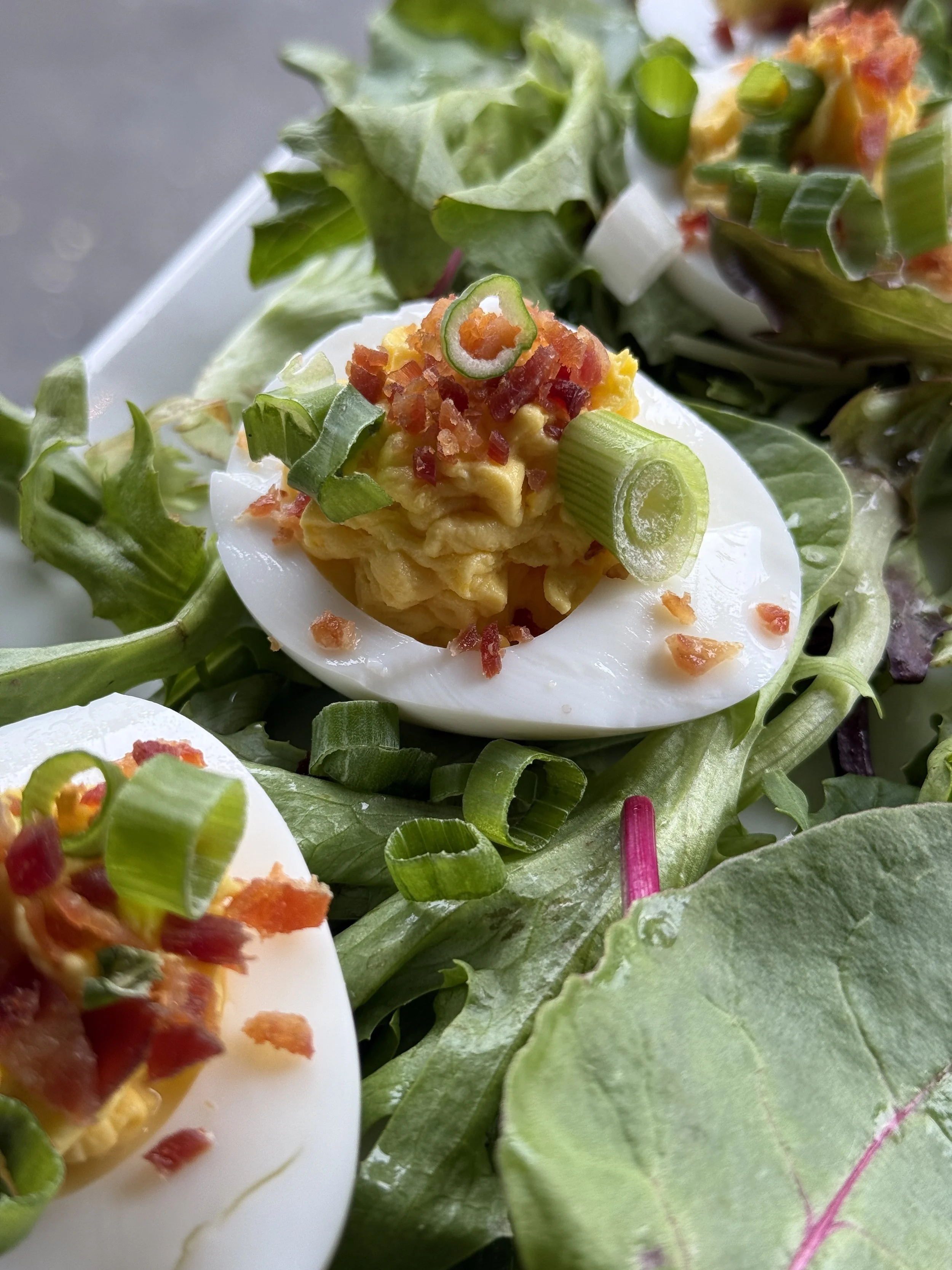 Close-up of a deviled egg topped with chopped green and red peppers and chopped bacon, resting on a bed of leafy greens.