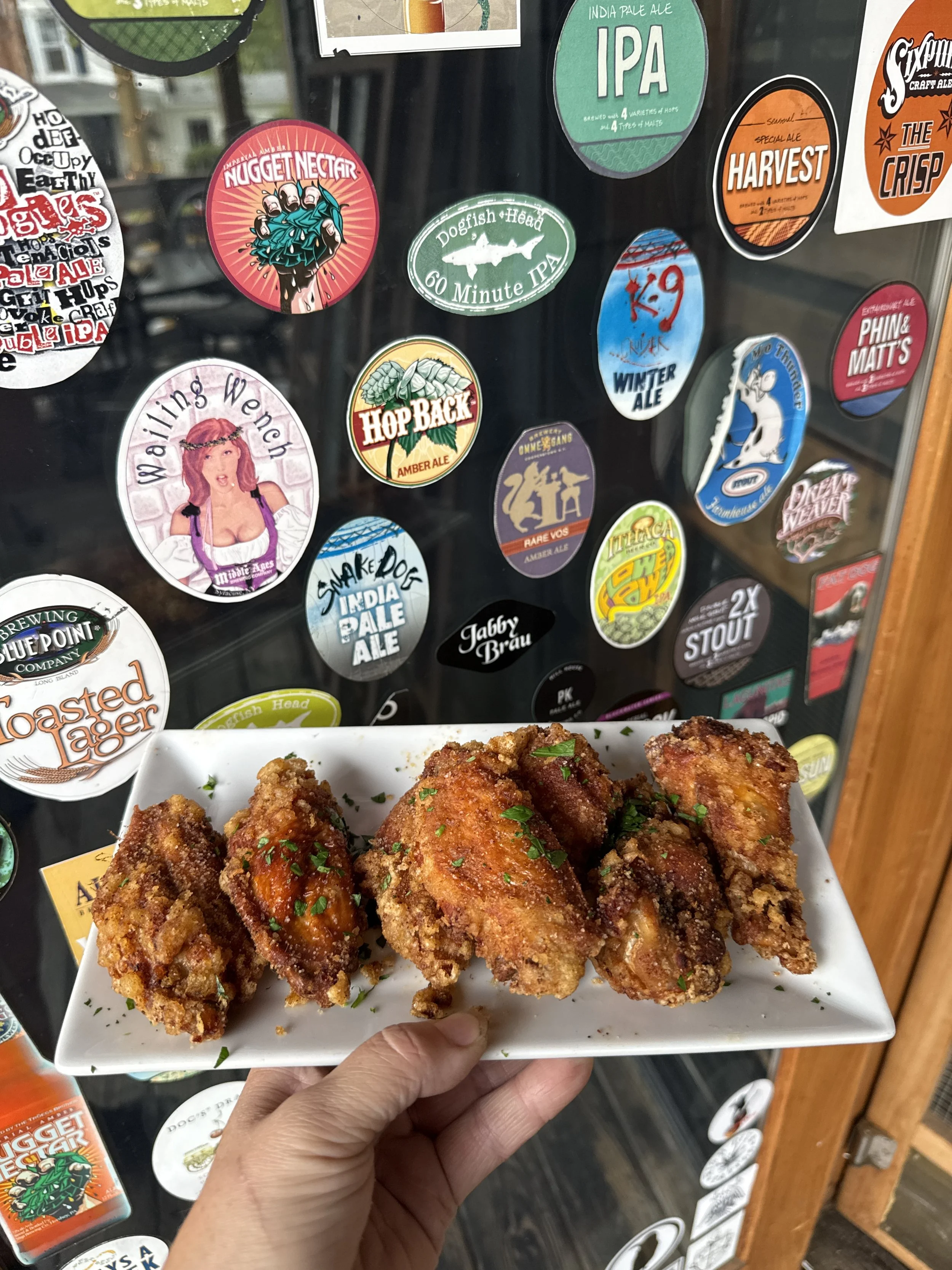 A hand holding a rectangular white plate with fried chicken drumsticks garnished with chopped herbs, in front of a glass wall covered with various colorful beer coasters.