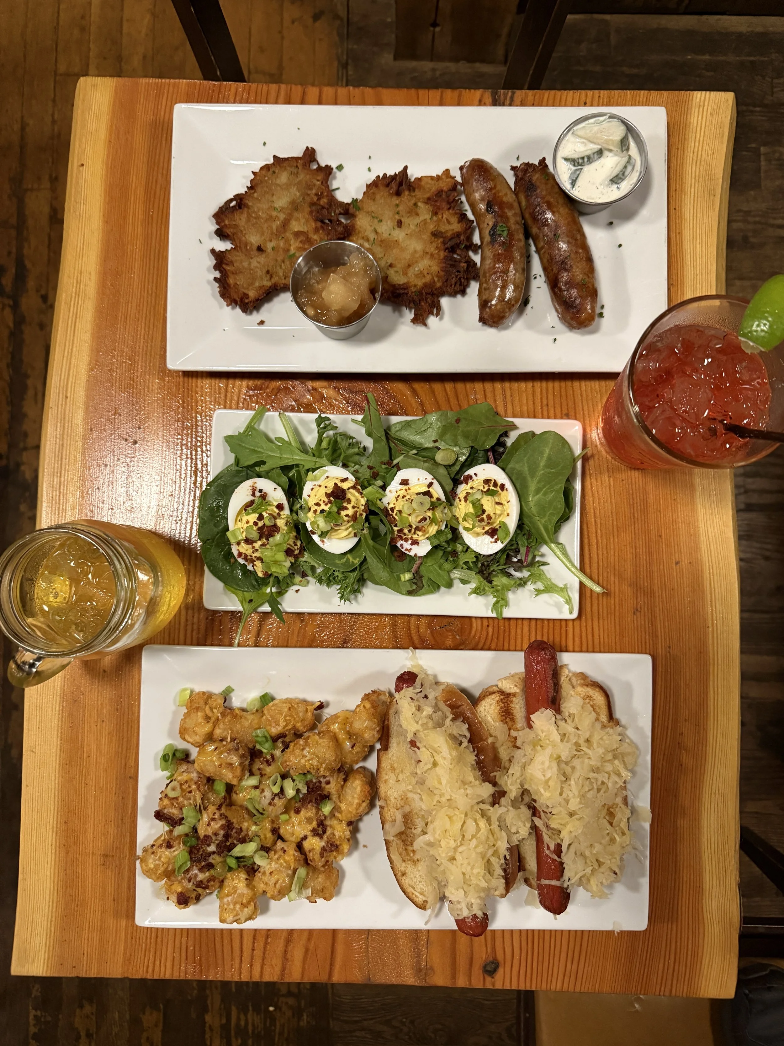 A wooden table with two rectangular white plates of food. The top plate has two hash brown potato cakes, two sausages, and small cups of sauces. The bottom plate has a salad with boiled eggs, a serving of chicken pieces, and a hot dog bun with grille