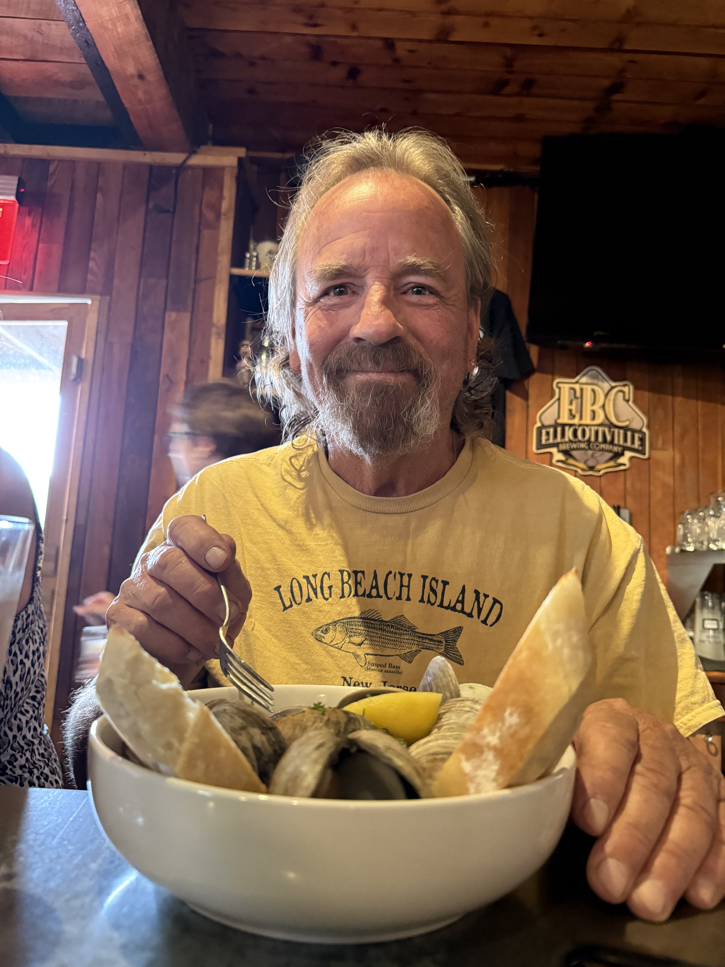 A man with gray hair and a beard, wearing a yellow shirt, sitting at a table with a seafood dish in front of him, inside a wooden-paneled restaurant or bar.