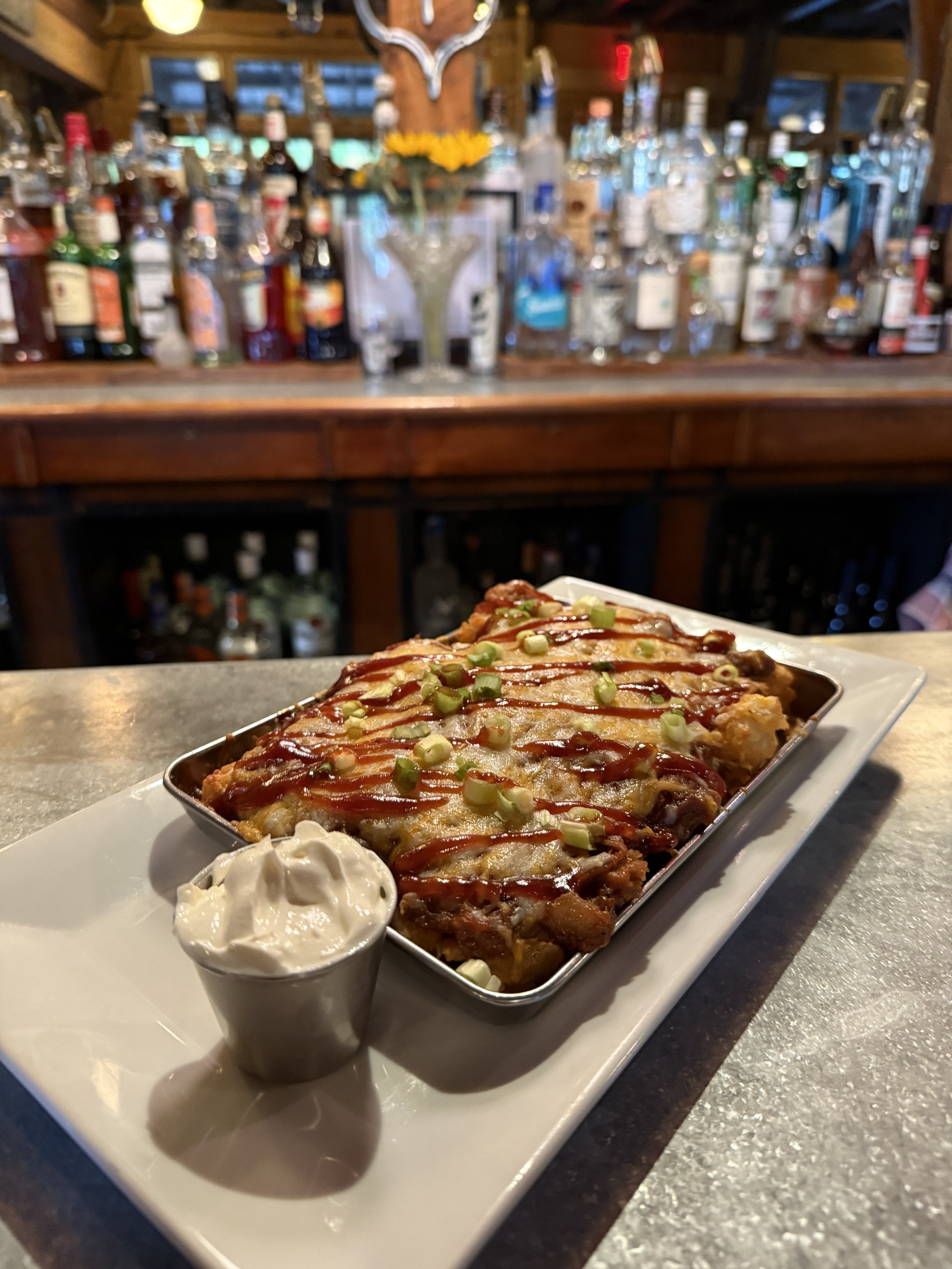Baked dish topped with green onions and sauce served with a side of sour cream, on a white rectangular plate in a restaurant setting with a bar in the background.