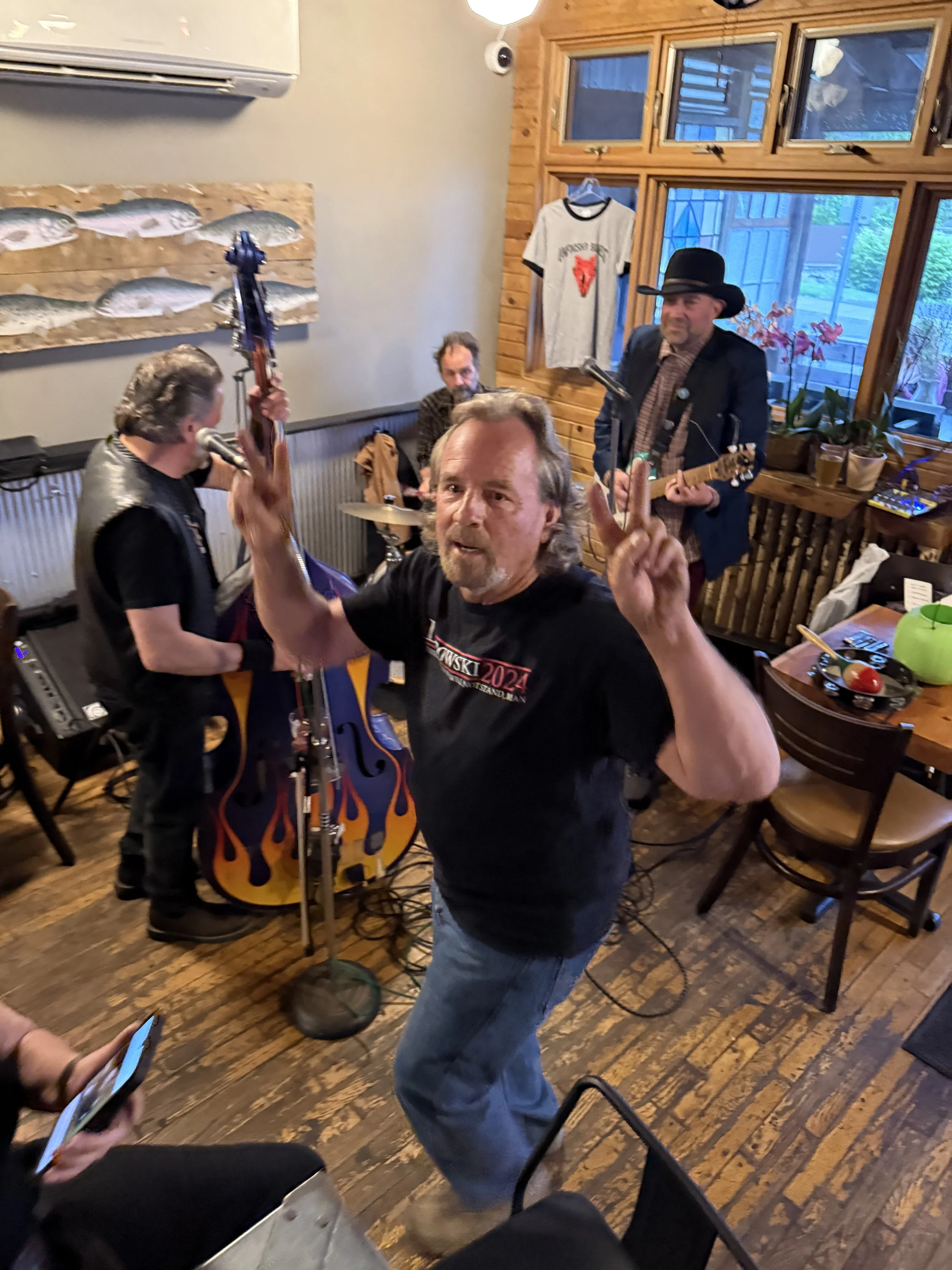 A man in a black T-shirt and jeans is dancing and making a peace sign. Behind him, three men are playing musical instruments including a guitar and drums. The setting appears to be a cozy wooden restaurant or cafe with large windows, a menu board, and various decorations including a t-shirt hanging on the wall and a painting of fish.